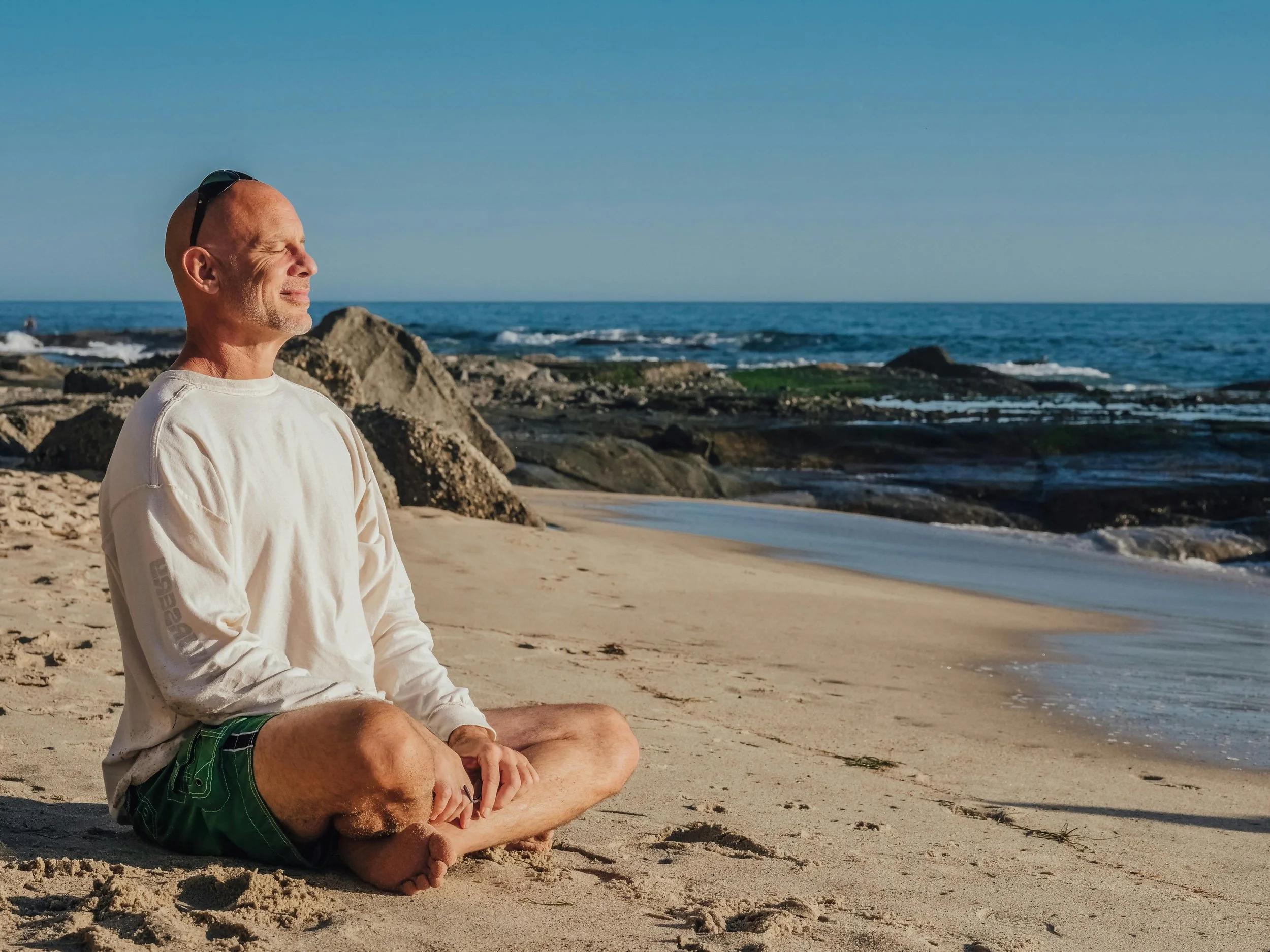 A man sitting cross-legged on the sandy beach, meditating with eyes closed, wearing a long sleeve shirt and green shorts, overlooking the ocean with rocks and waves in the background during daytime.