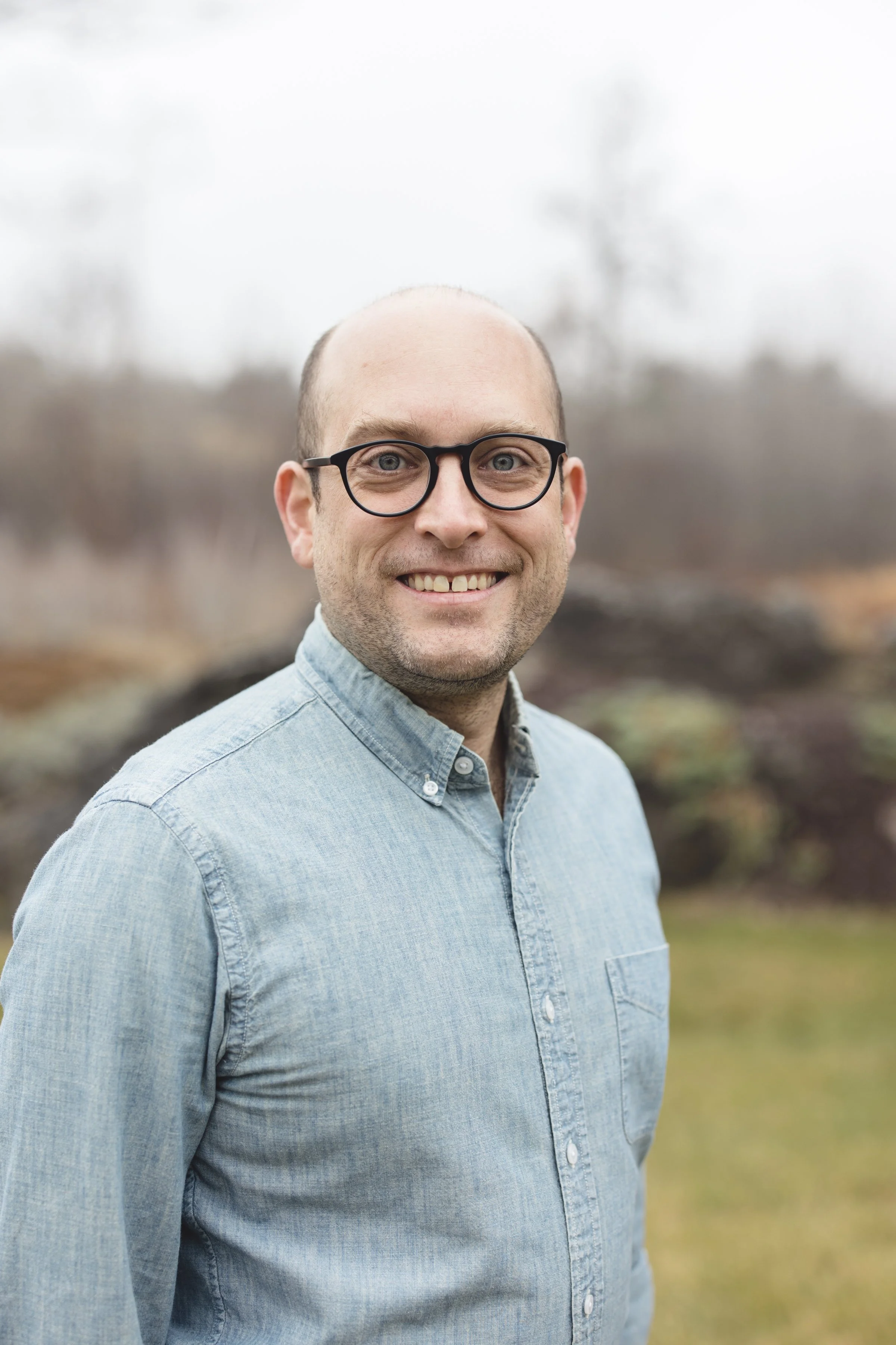 A man outdoors with a smile, wearing glasses and a light blue denim button-up shirt, with a blurred Natural background.
