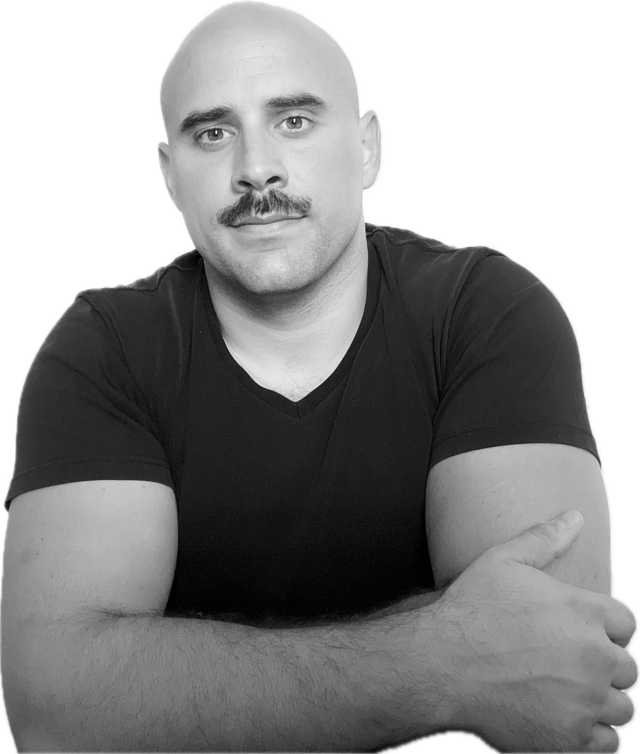 A black and white portrait of a man with a shaved head, mustache, wearing a black V-neck T-shirt, sitting with arms crossed.