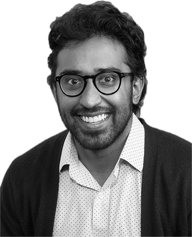 Headshot of a smiling man with glasses, dark curly hair, beard, wearing a light patterned shirt and dark cardigan.