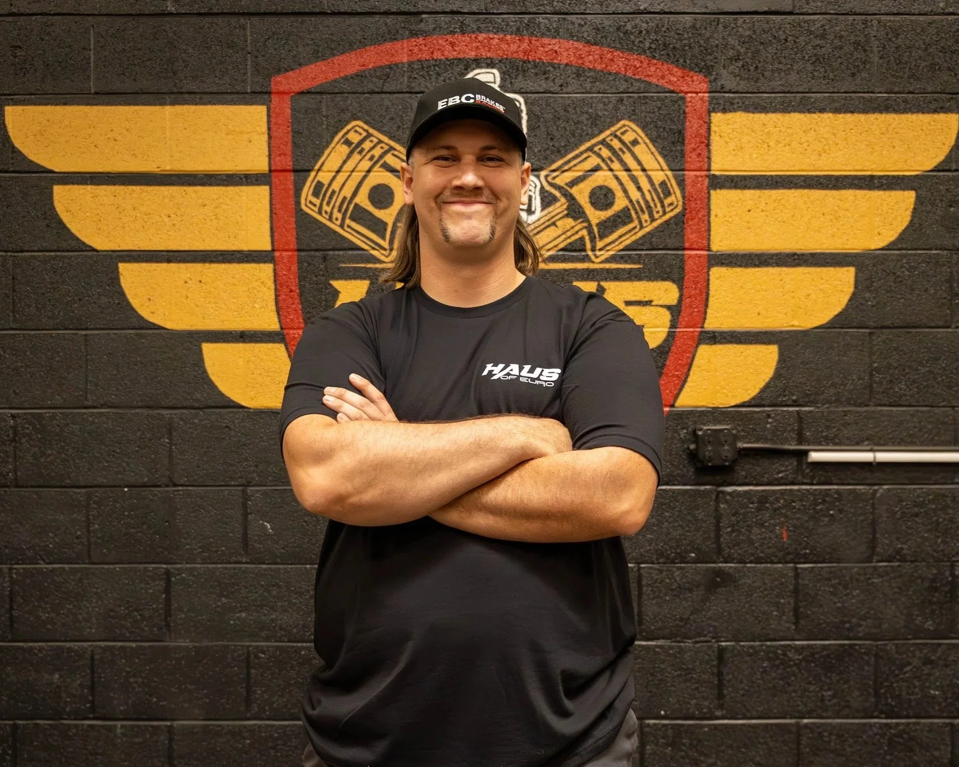 Man with arms crossed, smiling, wearing a black cap and black t-shirt, standing in front of a wall with a logo featuring winged dollar signs and a shield.