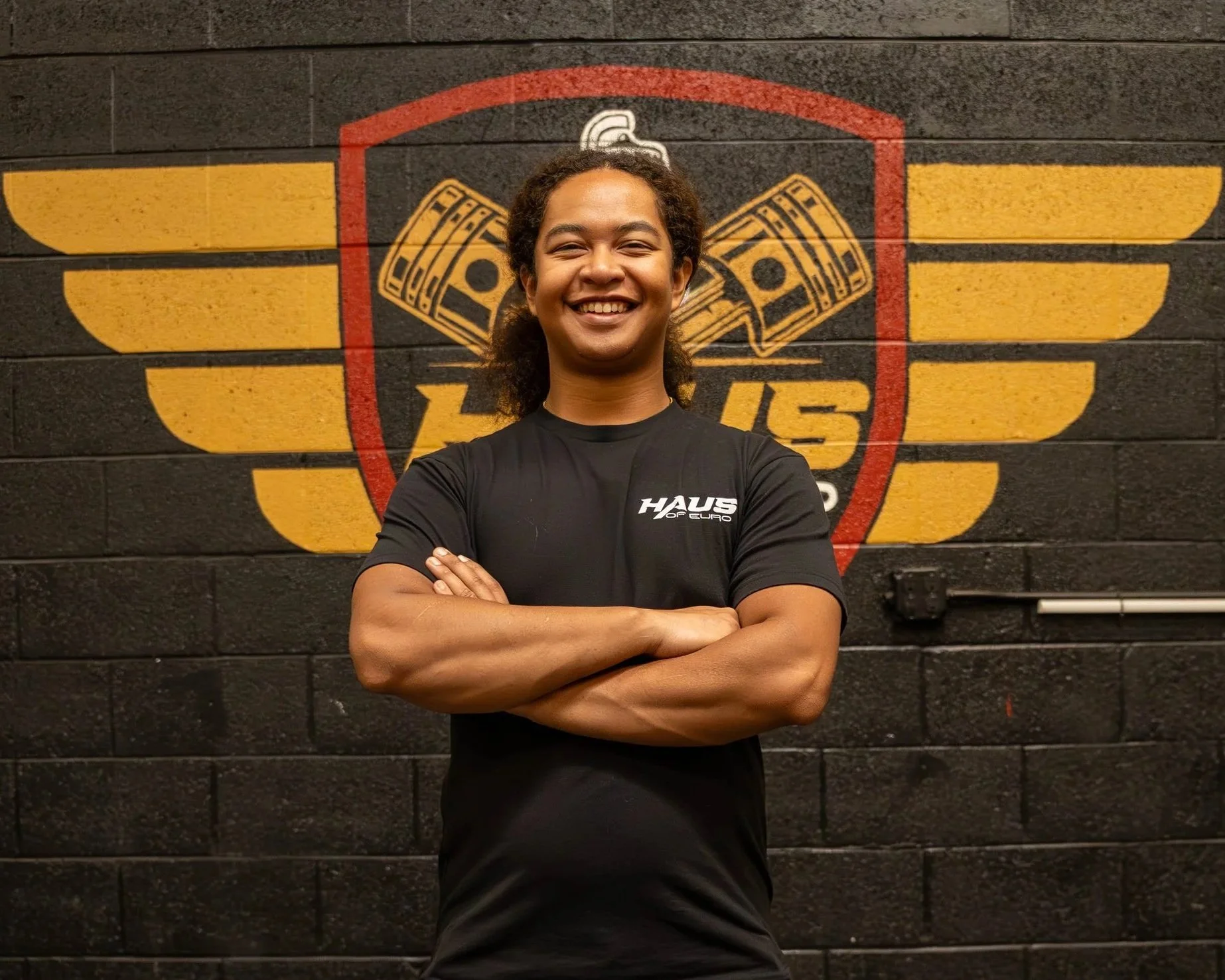 A young man with curly hair, smiling and standing with arms crossed in front of a black brick wall with a yellow and red logo featuring wings and money symbols.