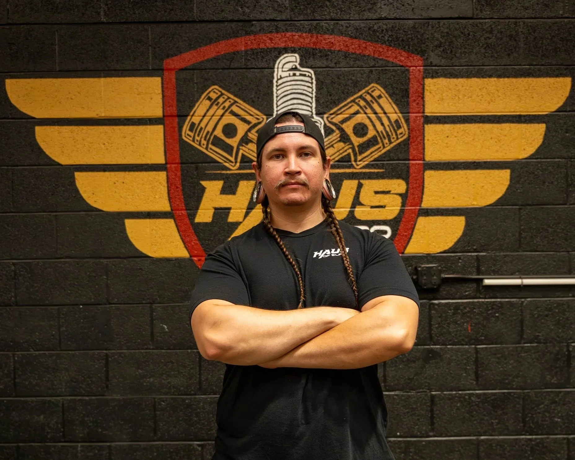 A man with long braided hair and earrings standing with arms crossed in front of a black wall with a logo that includes yellow wings, a syringe, money, and the words "HATES".