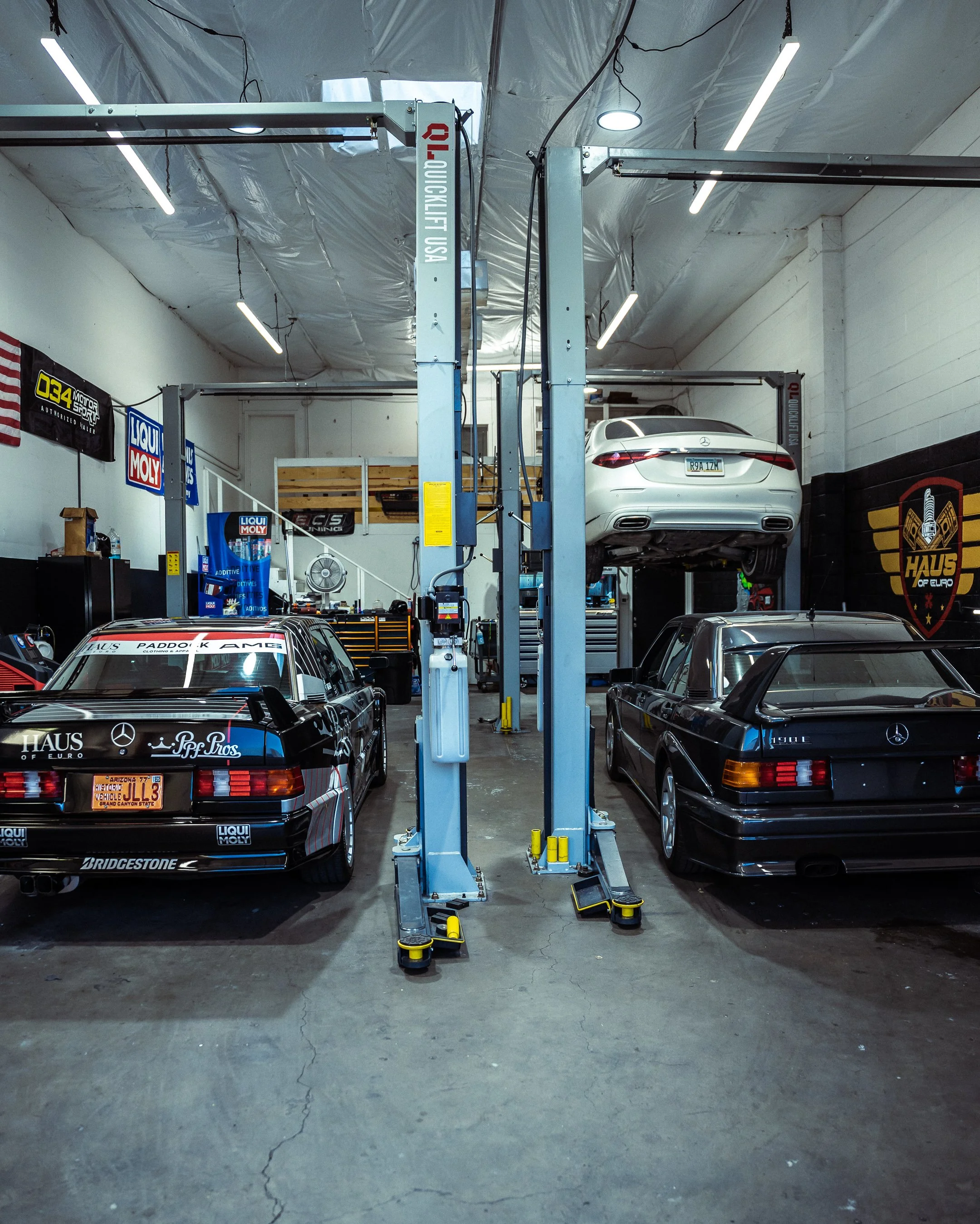 Two dark grey Mercedes-Benz 190 E models and a white E-Class on a lift receiving expert service at the Haus of Euro repair shop in Tempe, AZ.