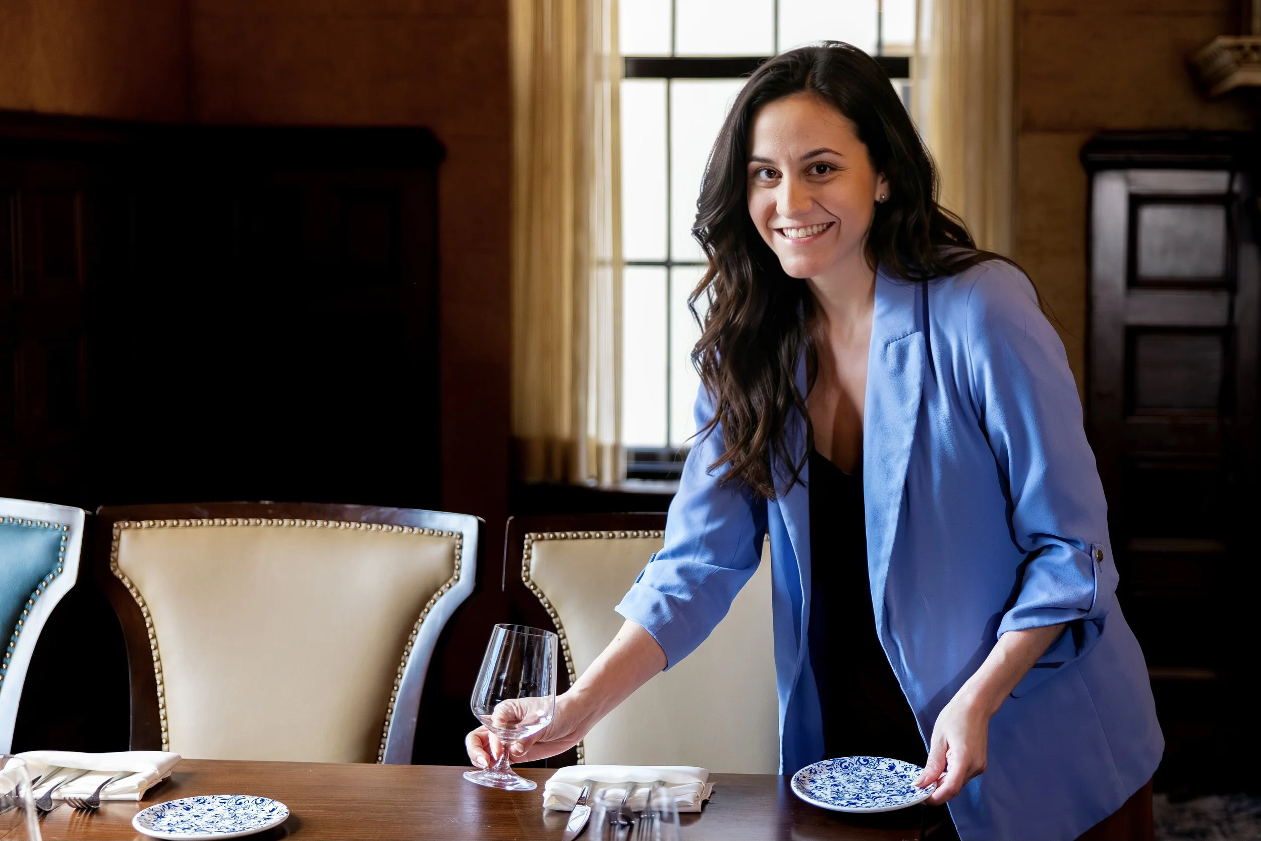 A woman with dark hair in a bun wearing a white button-up shirt and yellow pants standing in a kitchen with a smile.