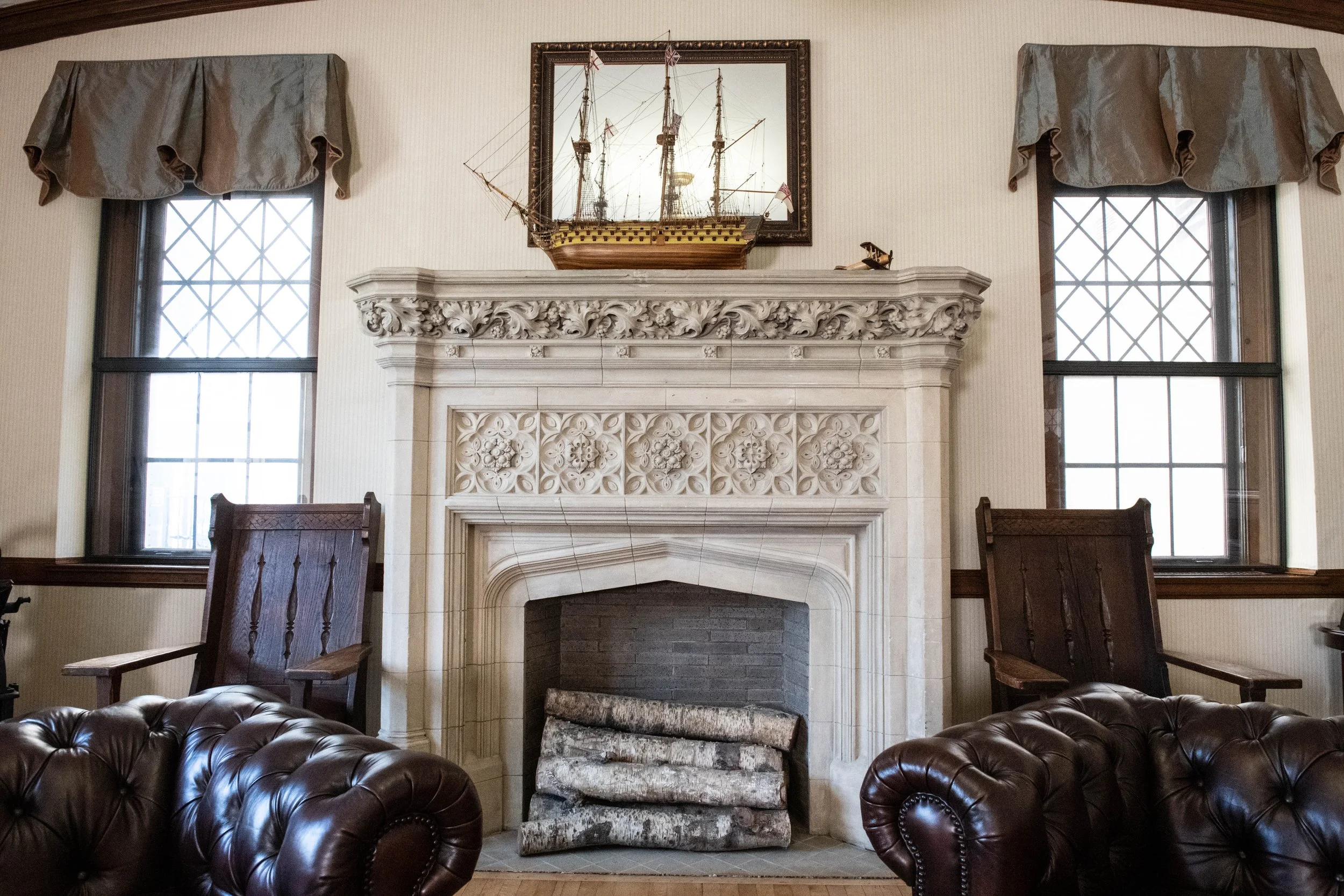 Living room with a carved stone fireplace, two wooden chairs, leather armchairs, two windows with curtains, a model ship, and framed artwork.