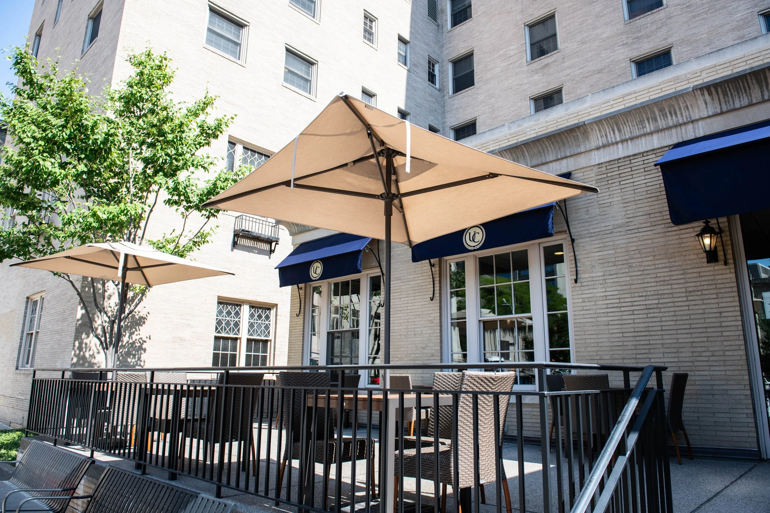 Outdoor patio area with wicker chairs and tables, beige umbrellas, and a black metal railing, outside a multi-story brick building with large windows and blue awnings.
