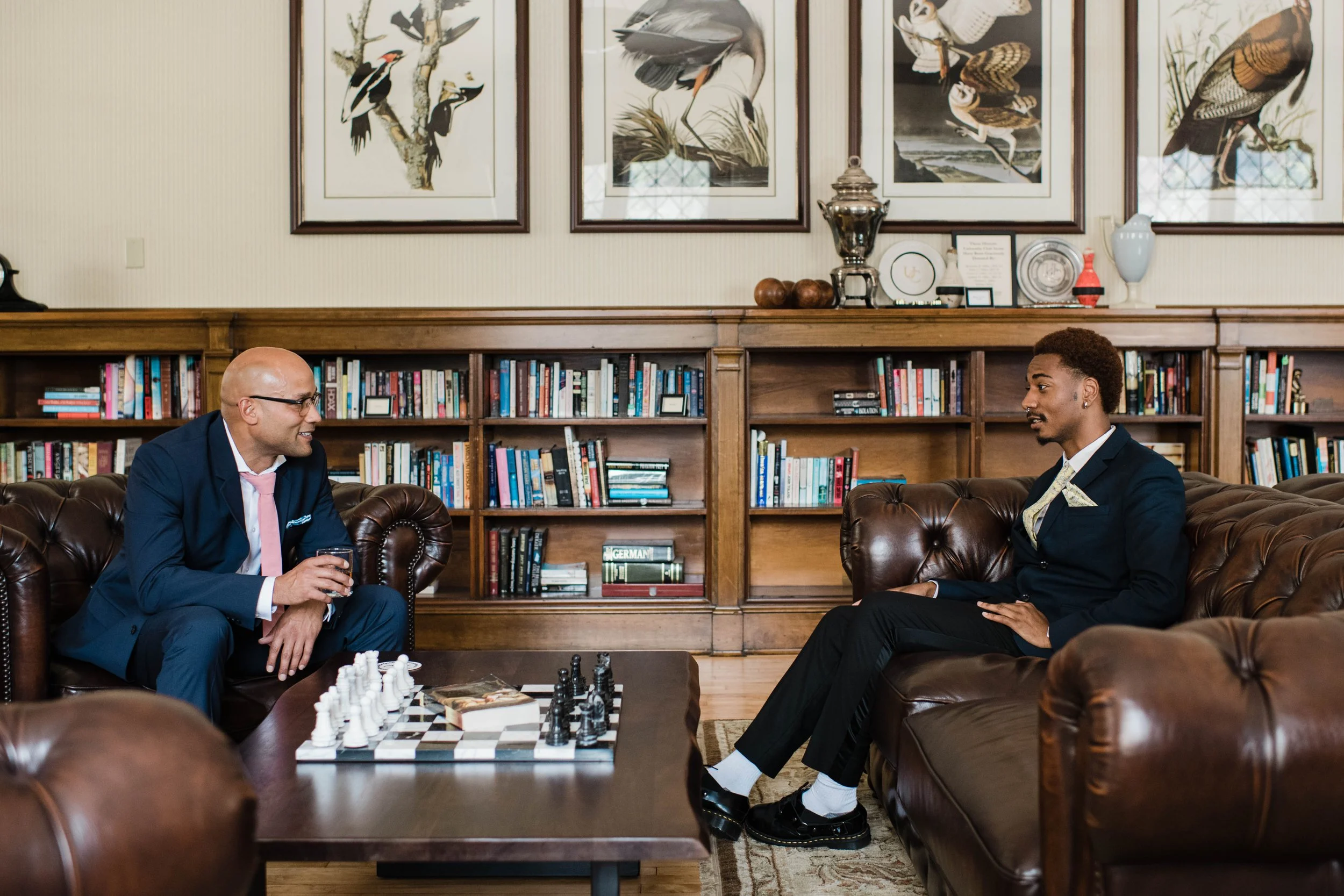 Two men in suits playing chess on a coffee table in a library or office, with framed bird artwork, books, and decorative items on shelves behind them.