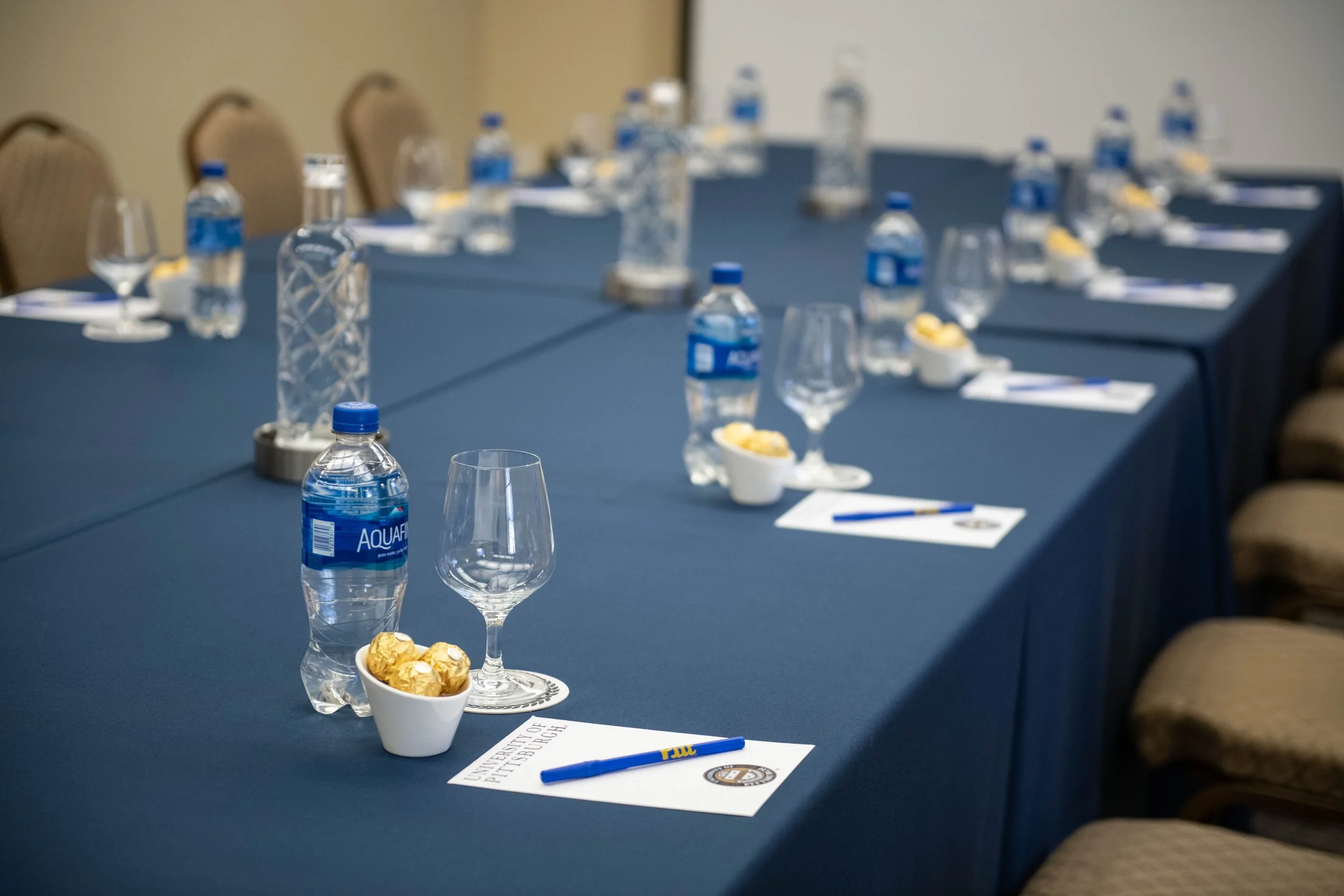 Conference room table set with bottled water, wine glasses, small dishes of snacks, and writing materials, with chairs around the table.