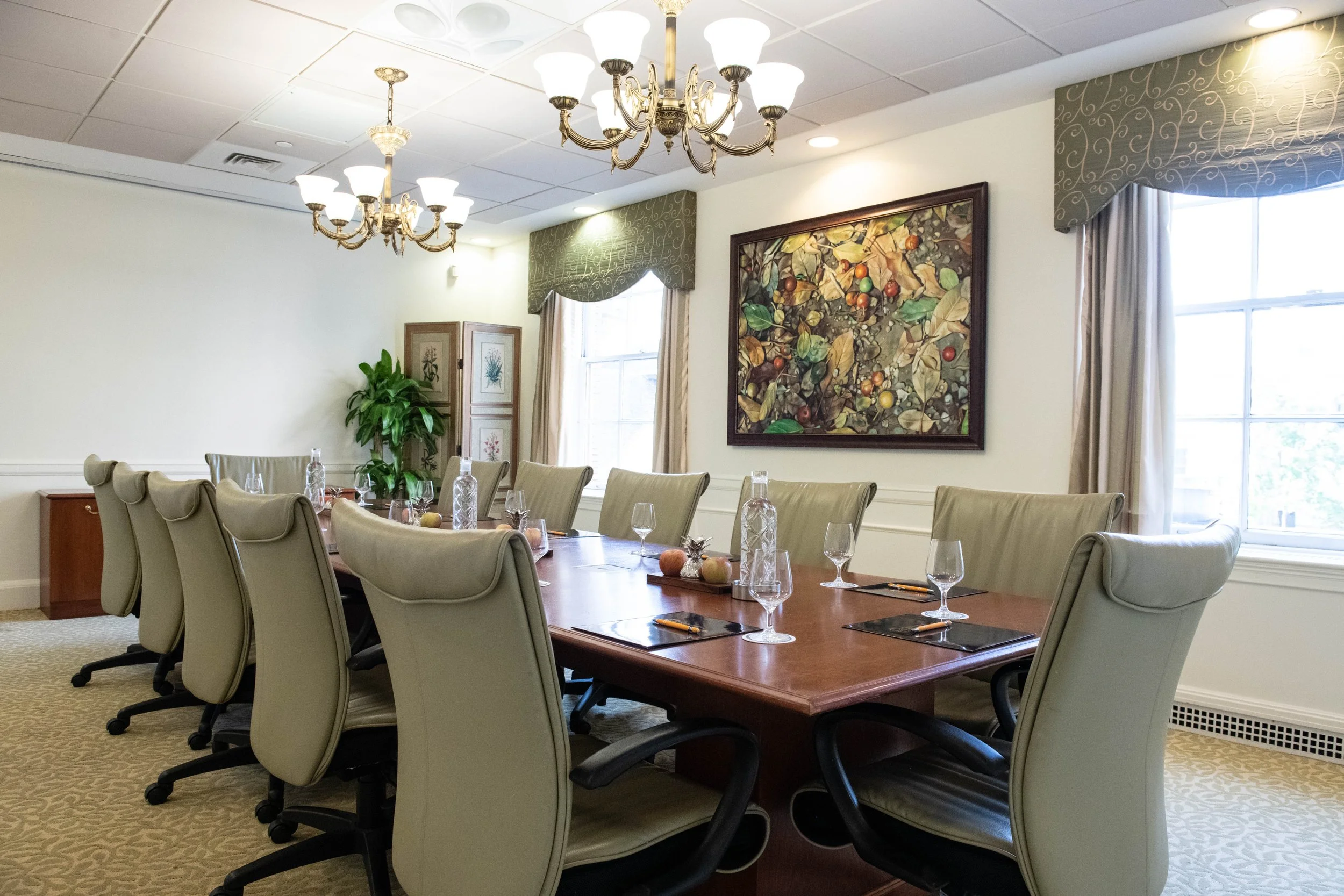 A conference room with a long wooden table surrounded by beige upholstered office chairs. The table is set with water bottles, glasses, and notepads. There is a painting of leaves and fruit on the wall, and the room has large windows with curtains, c