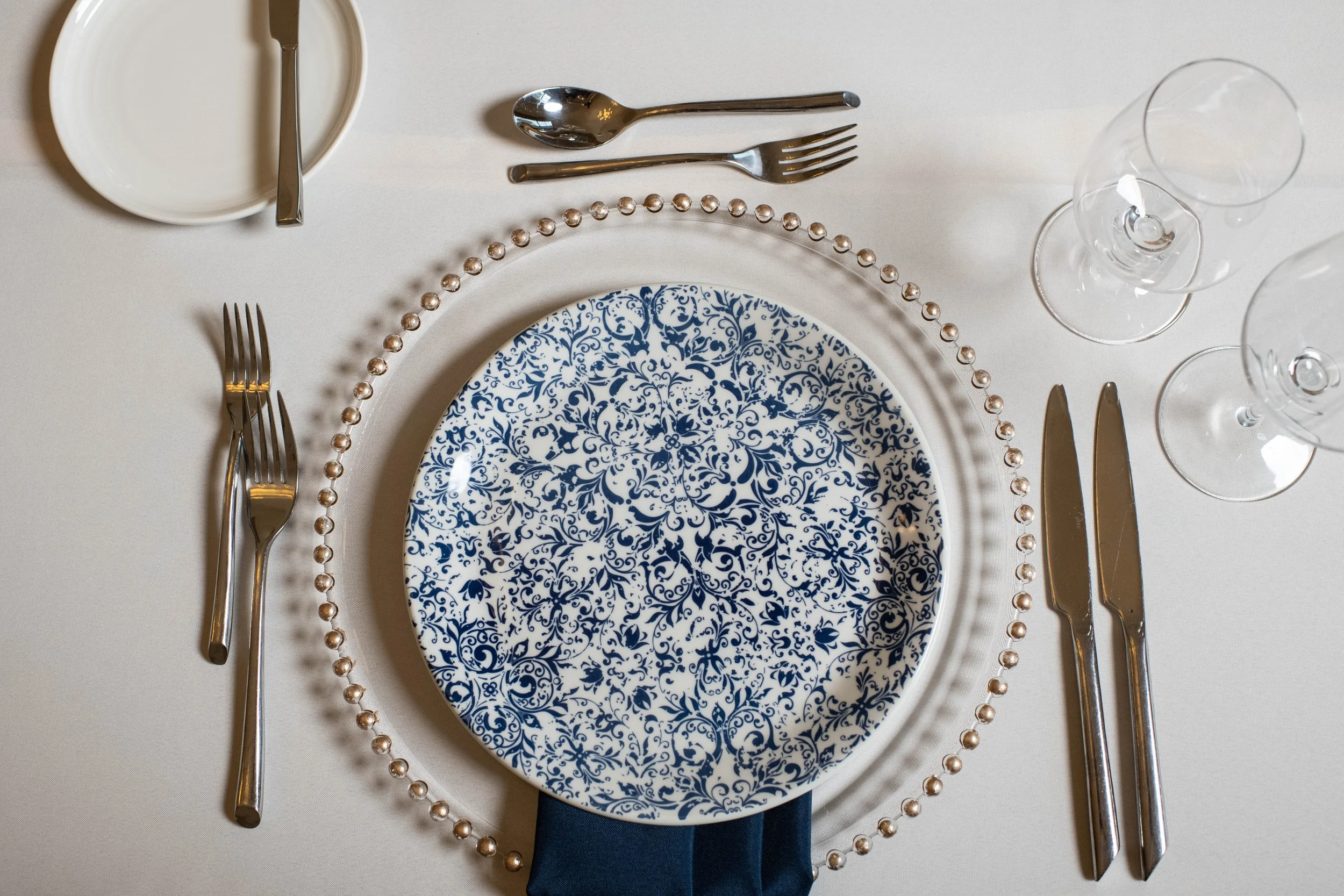A formal table setting with a blue and white patterned plate, pearl-bordered glass charger, stainless steel utensils, two empty wine glasses, and a small bowl with a spoon on a white tablecloth.