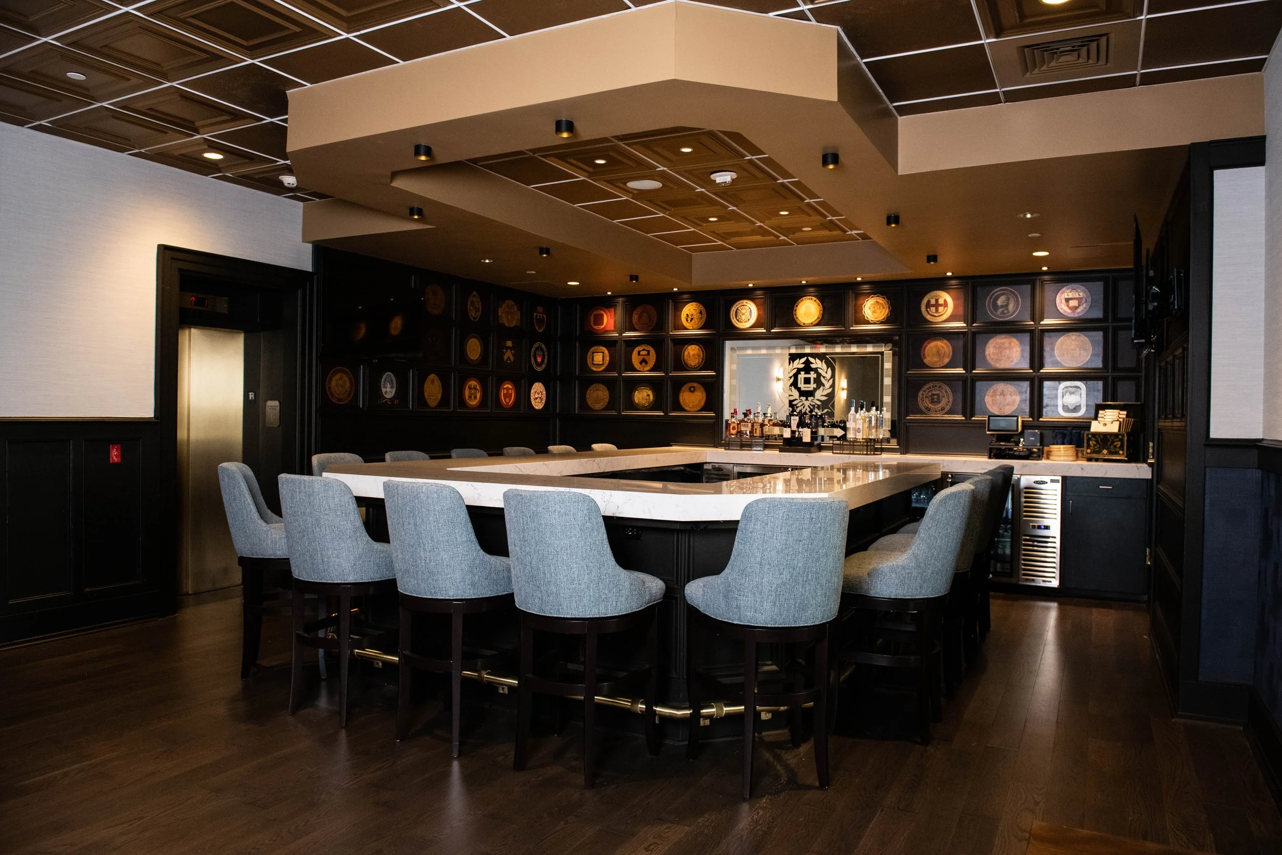 College Room at the University Club at Pitt | An elegant bar with a U-shaped white marble countertop bar, light gray upholstered chairs, black wooden flooring, and a black wall decorated with framed shields and plaques. Open to the public M-F