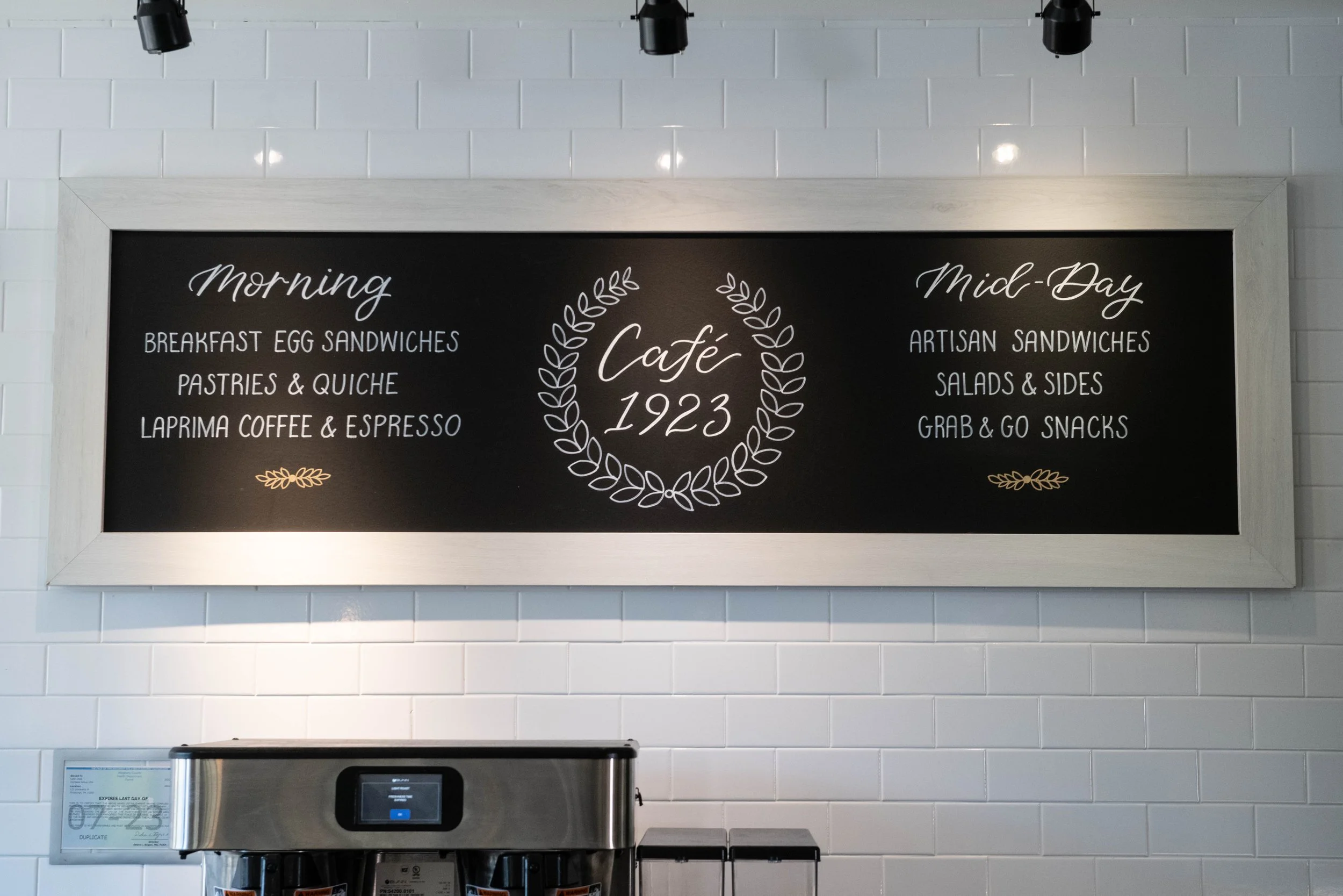 Black chalkboard menu with white and gold writing at Café 1923, split into morning and mid-day sections listing food offerings, with a coffee machine below.