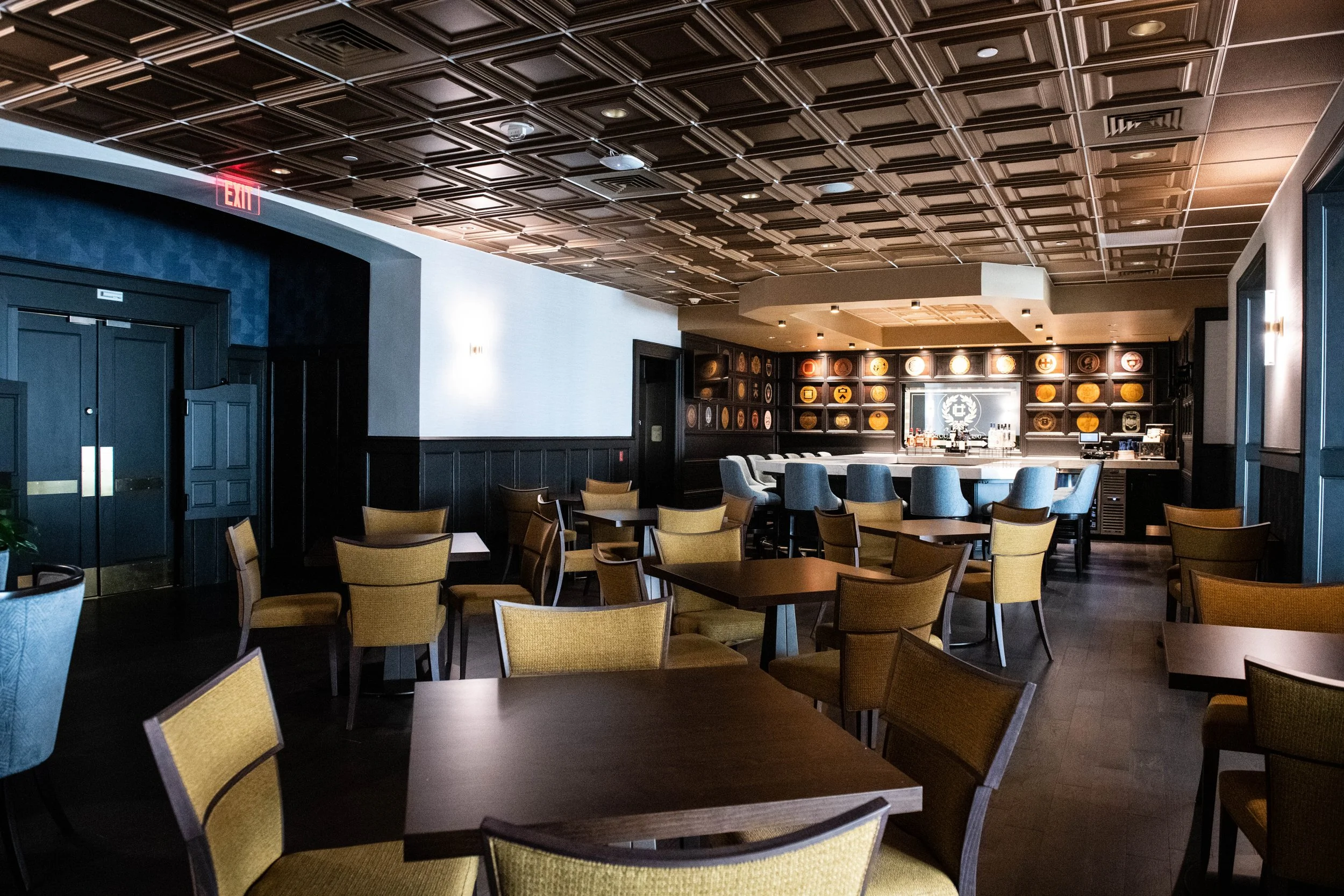 College Room at the University Club at Pitt | An empty hotel bar with wooden tables and chairs, a dark wood-paneled ceiling, and a well-lit bar area with stools and decorative wall art. Open to the public Monday - Friday.