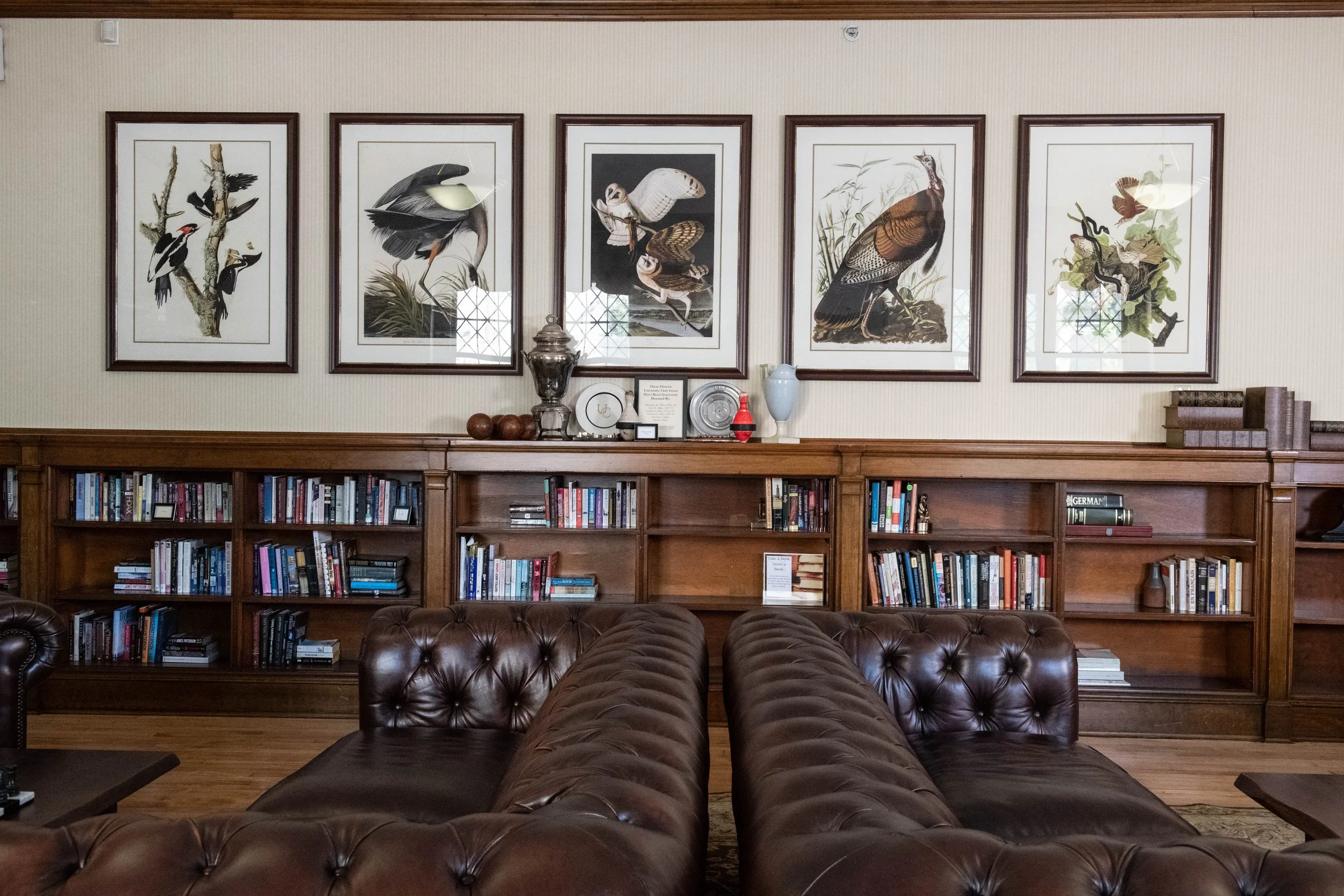 A living room with a wooden bookshelf, leather couches, and framed bird prints on the wall.