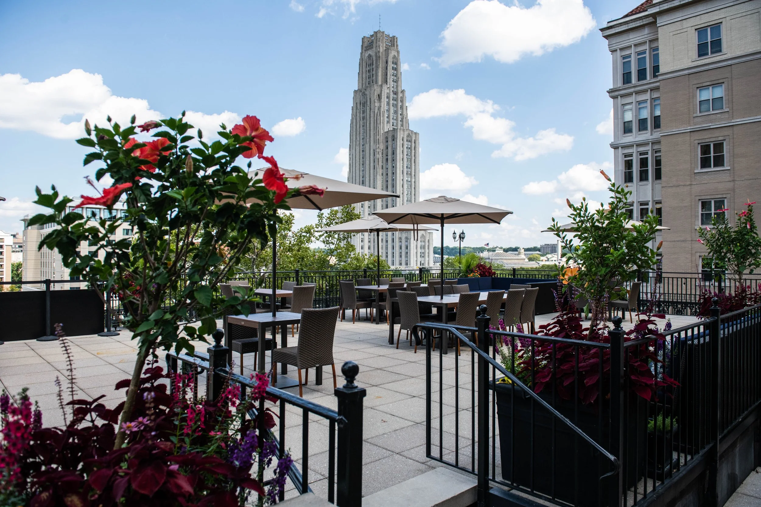 A rooftop cafe with tables, chairs, and umbrellas under a partly cloudy sky, with a tall art deco building and cityscape in the background, adorned with potted plants and flowers.