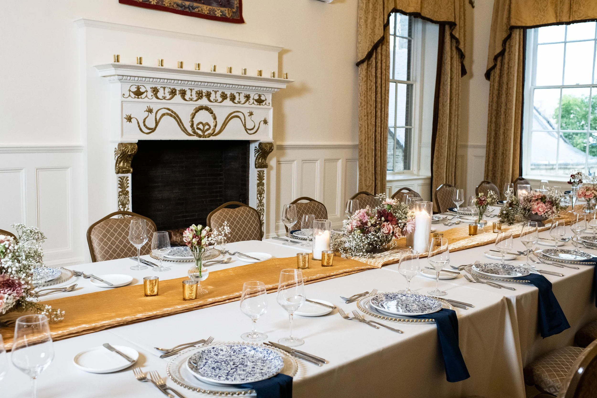 Decorated dining table with flowers, candles, and dinnerware in a room with large windows and a fireplace.