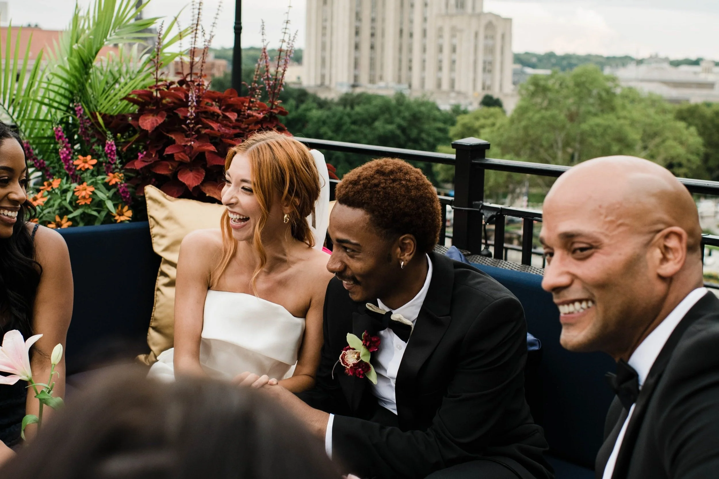 People wearing formal attire enjoying a conversation on a balcony during a celebration, with a cityscape and greenery in the background.