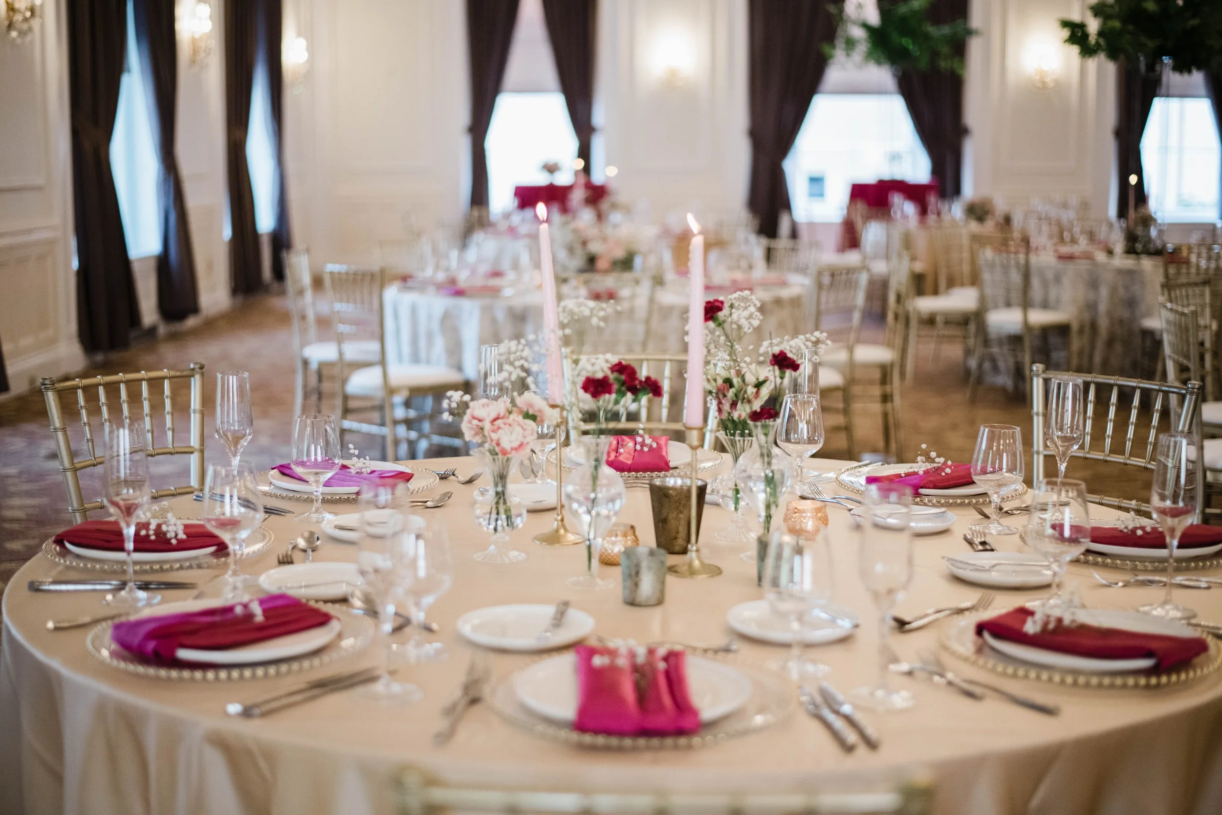 Ballroom B at the University Club | Elegant banquet table set with pink napkins, glassware, plates, and floral centerpieces in a decorated room with curtains and chandeliers.