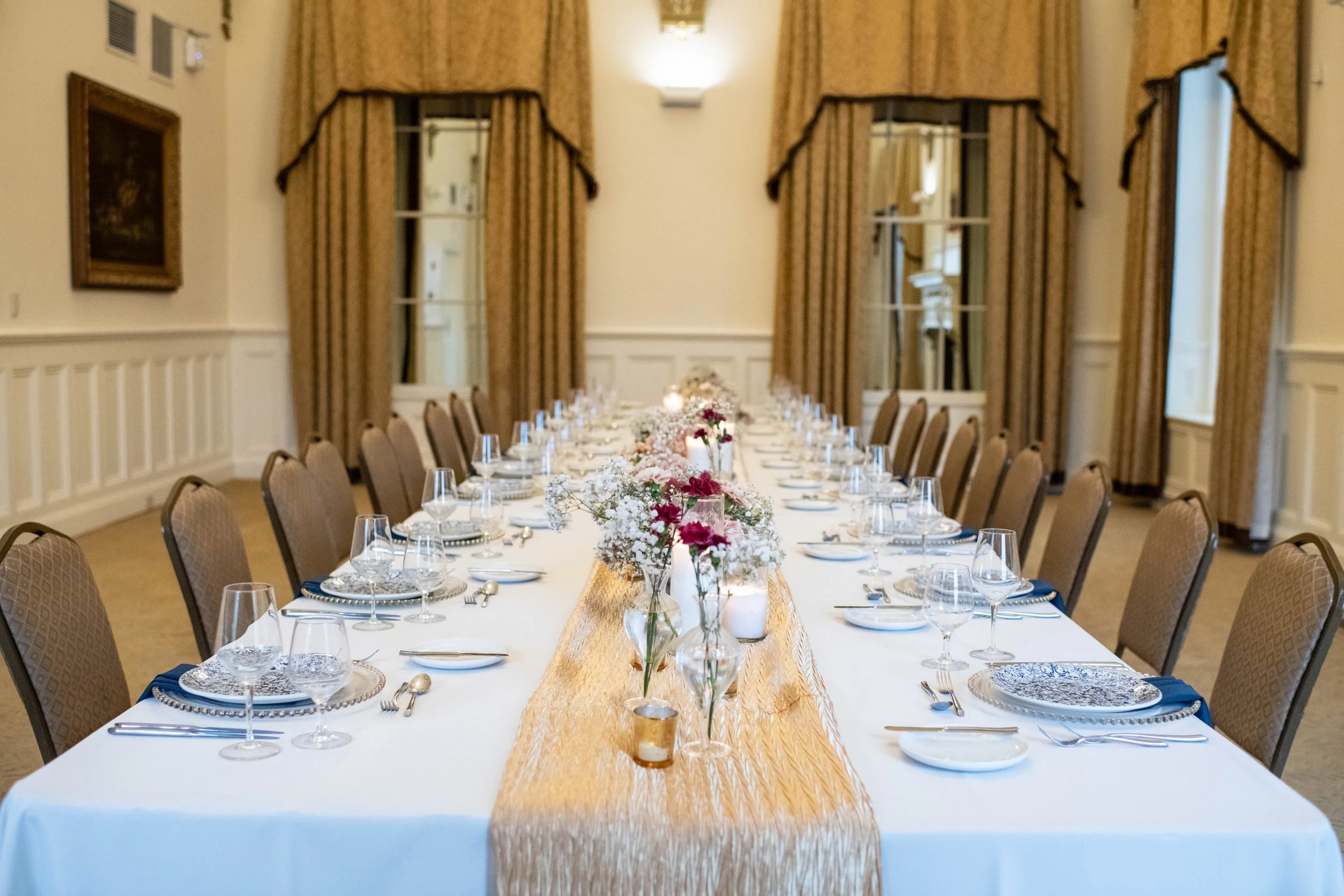 A long rectangular dining table set for a formal event, with a white tablecloth, floral centerpieces, and place settings including plates, glasses, and cutlery. The room has beige walls, large windows with gold curtains, and warm lighting.