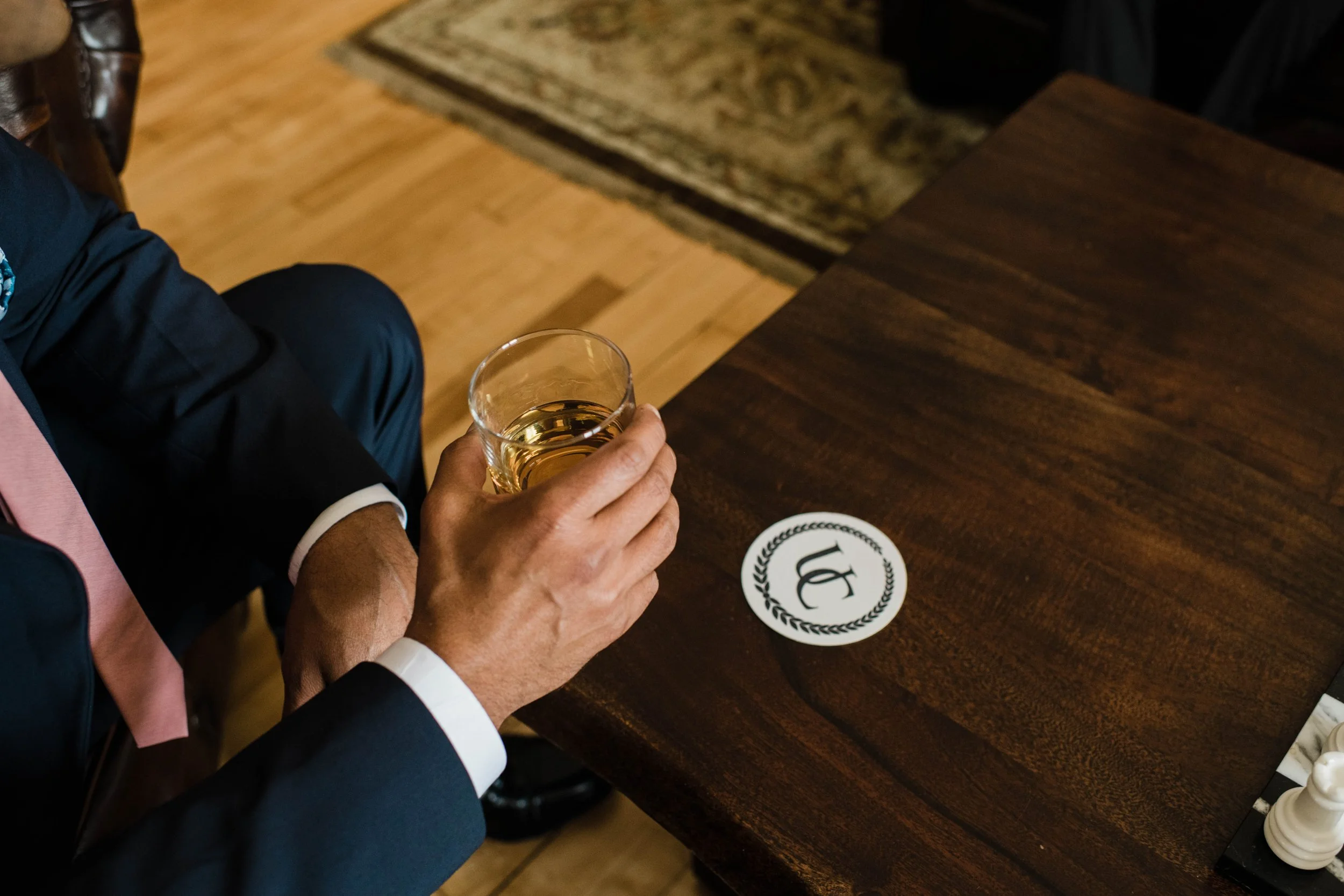 A man in a suit holding a glass of amber-colored beverage, sitting at a wooden table with a white circular coaster featuring a black 'C' logo.