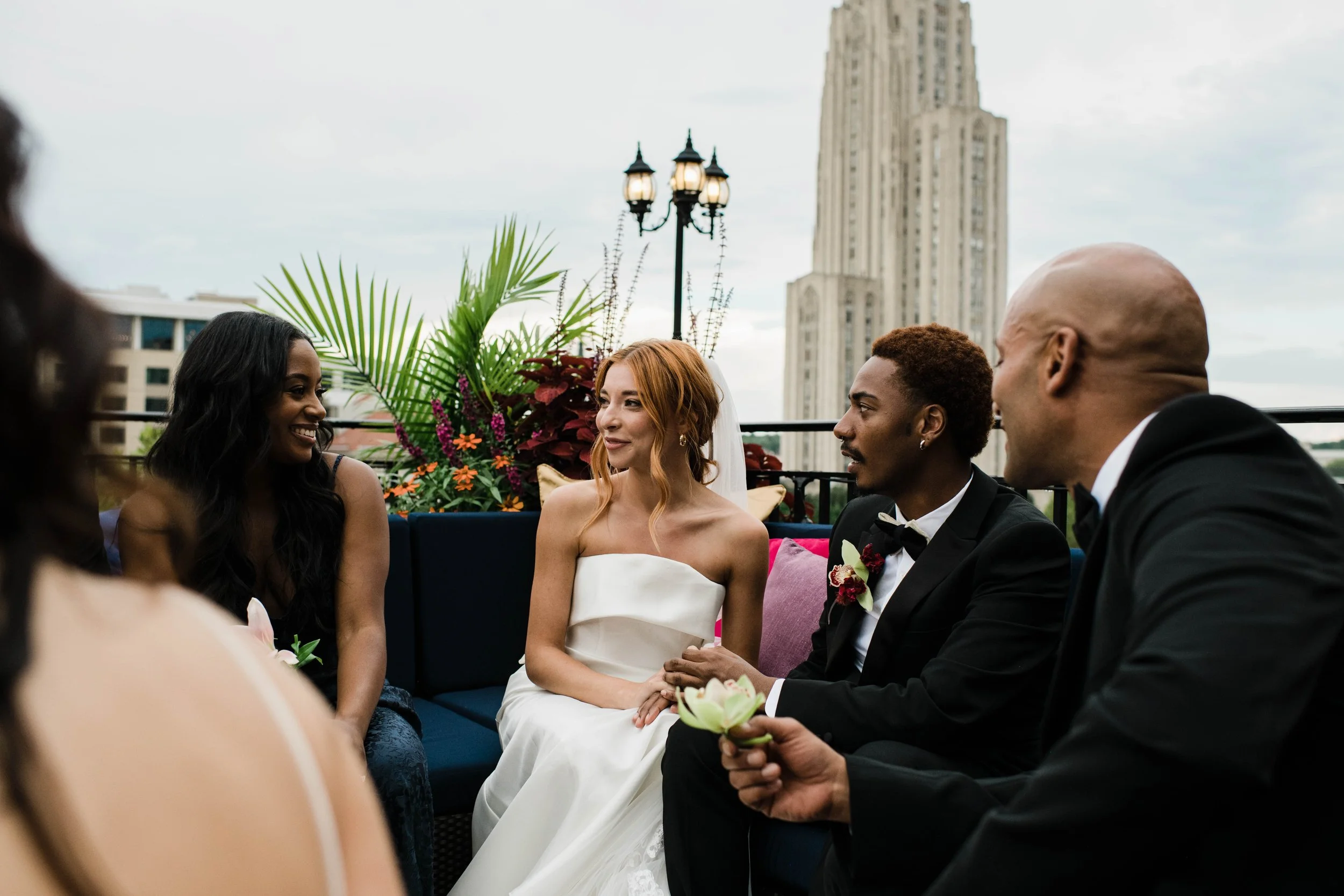 Terrace at the University Club | A group of people, including a bride in a white wedding dress and groom in a black tuxedo, sitting outside on a terrace with city buildings in the background, engaged in a conversation.