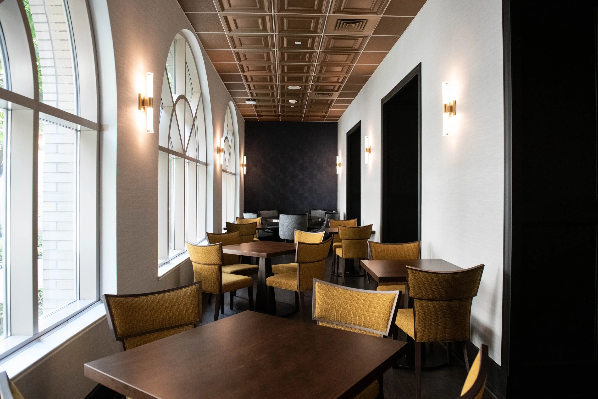 College Room at the University Club at Pitt | Interior view of a modern restaurant or cafe with large arched windows, yellow chairs, wooden tables, wall sconces lighting, with a dark accent wall, coffered ceiling. Open to the public Monday-Friday.