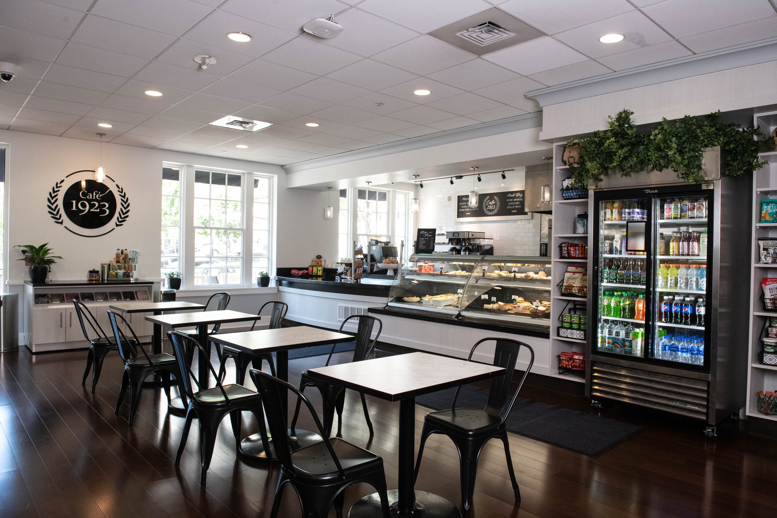 Cafe 1923 at University Club | Inside a modern cafe with white walls, dark wood floors, and black metal chairs. A display case with pastries, a beverage case, and a counter with coffee equipment are visible. Open to public Monday-Friday.