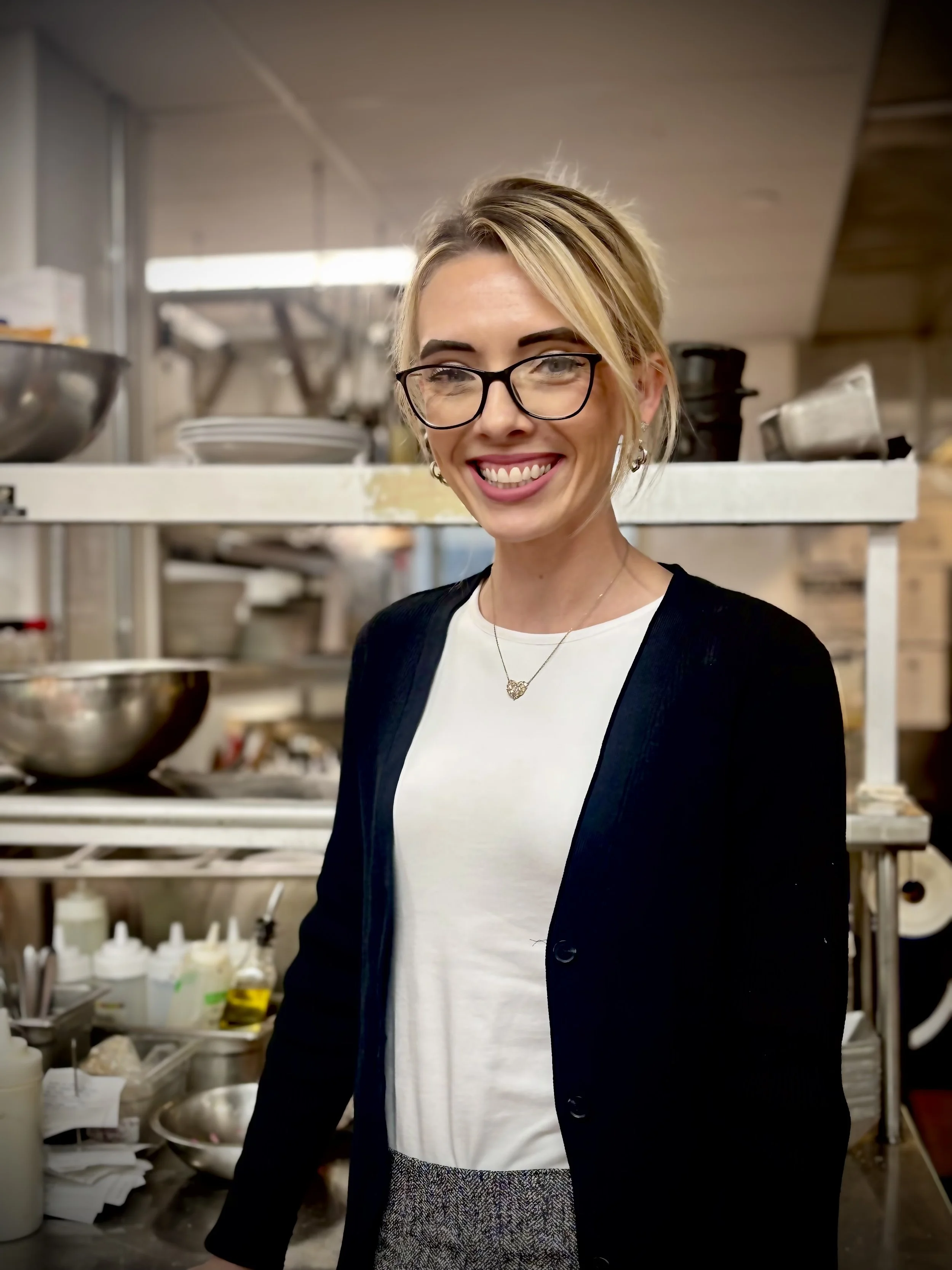 A woman with glasses and blonde hair smiling in a kitchen, wearing a black cardigan, white shirt, and a necklace, with kitchen utensils and bowls in the background.