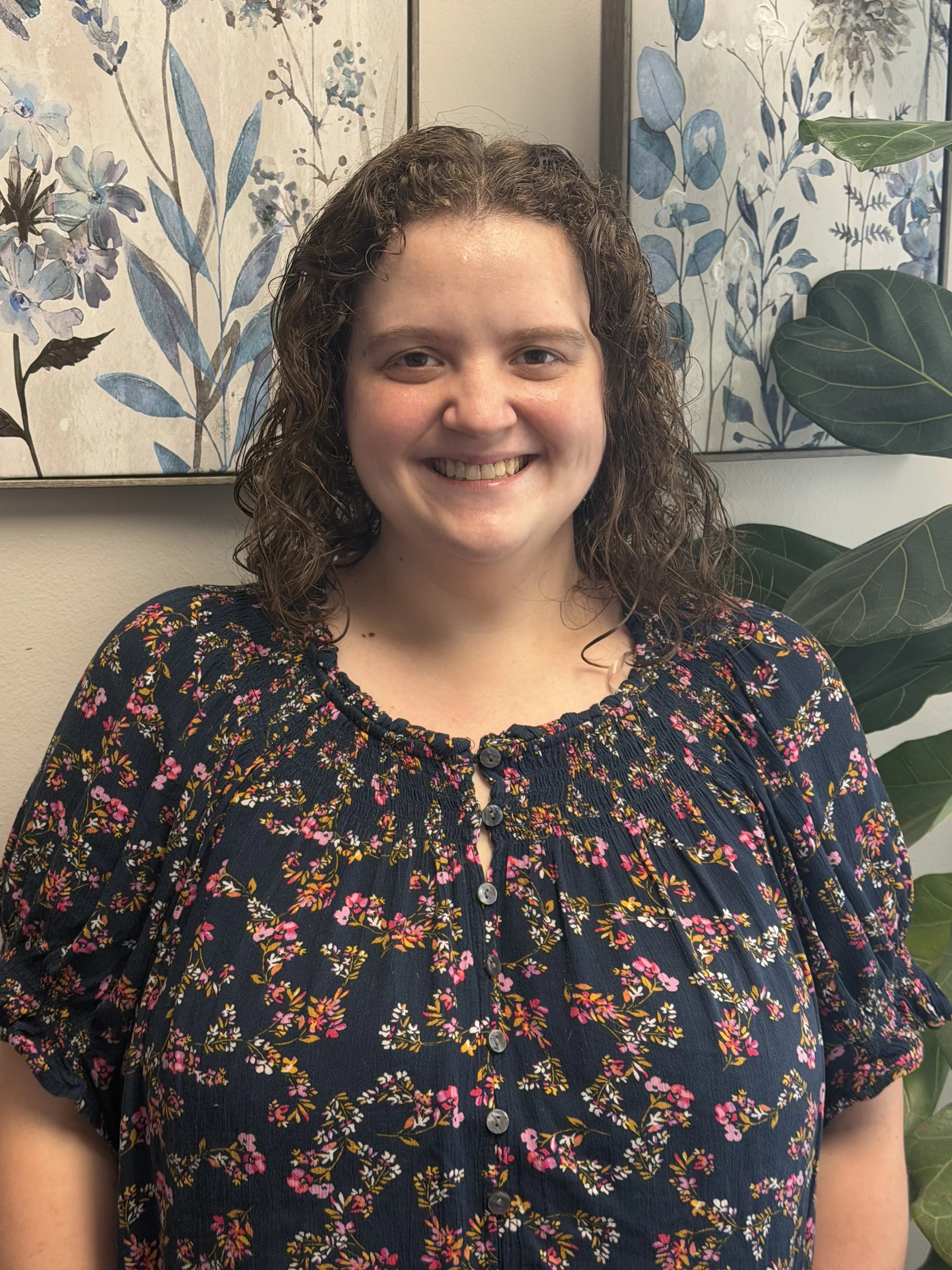 A woman with curly brown hair smiling in front of floral and leaf-patterned artwork and green plants.