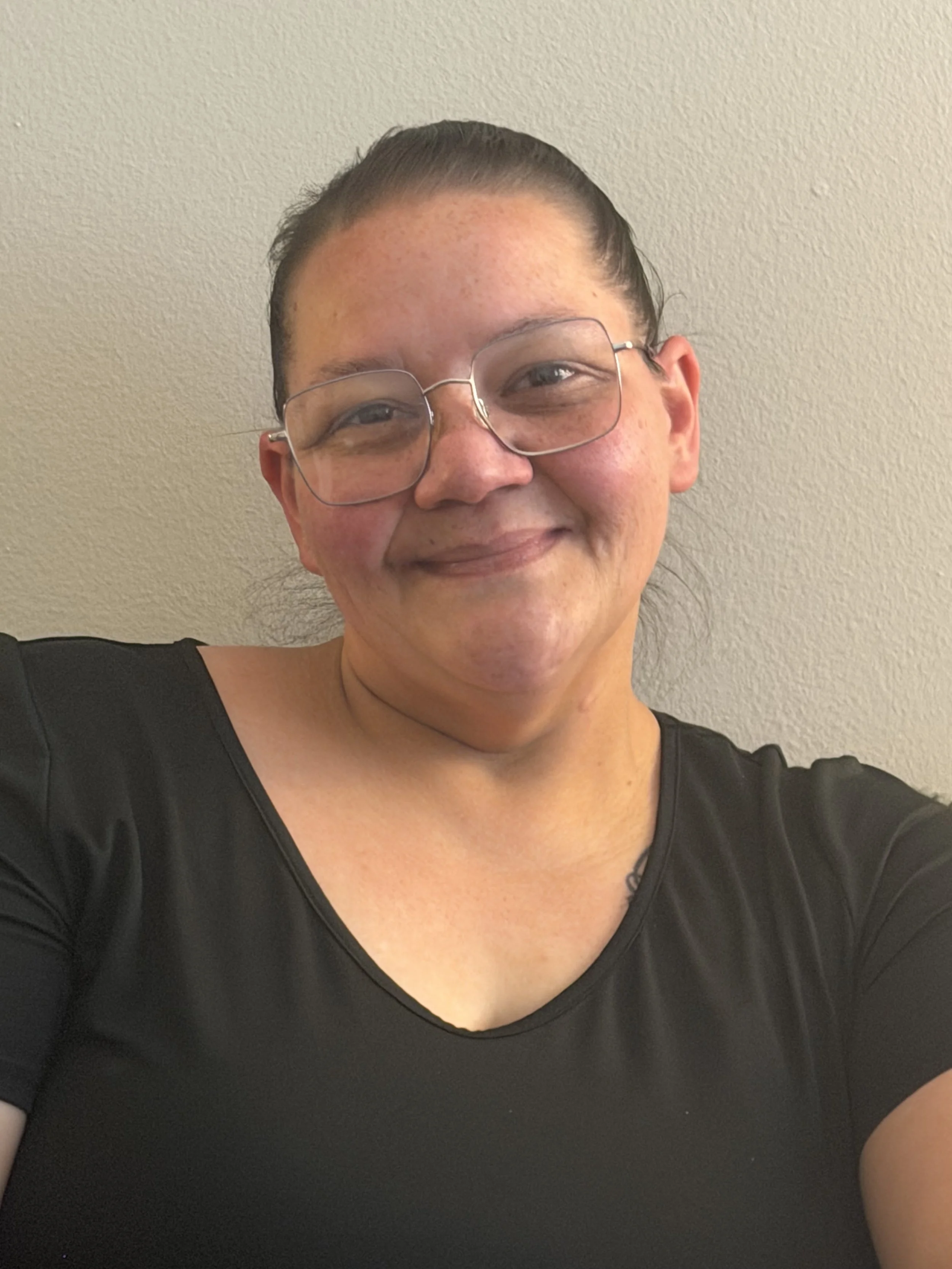 Close-up of a smiling woman with glasses, black shirt, and tied-back hair, against a plain light-colored wall.