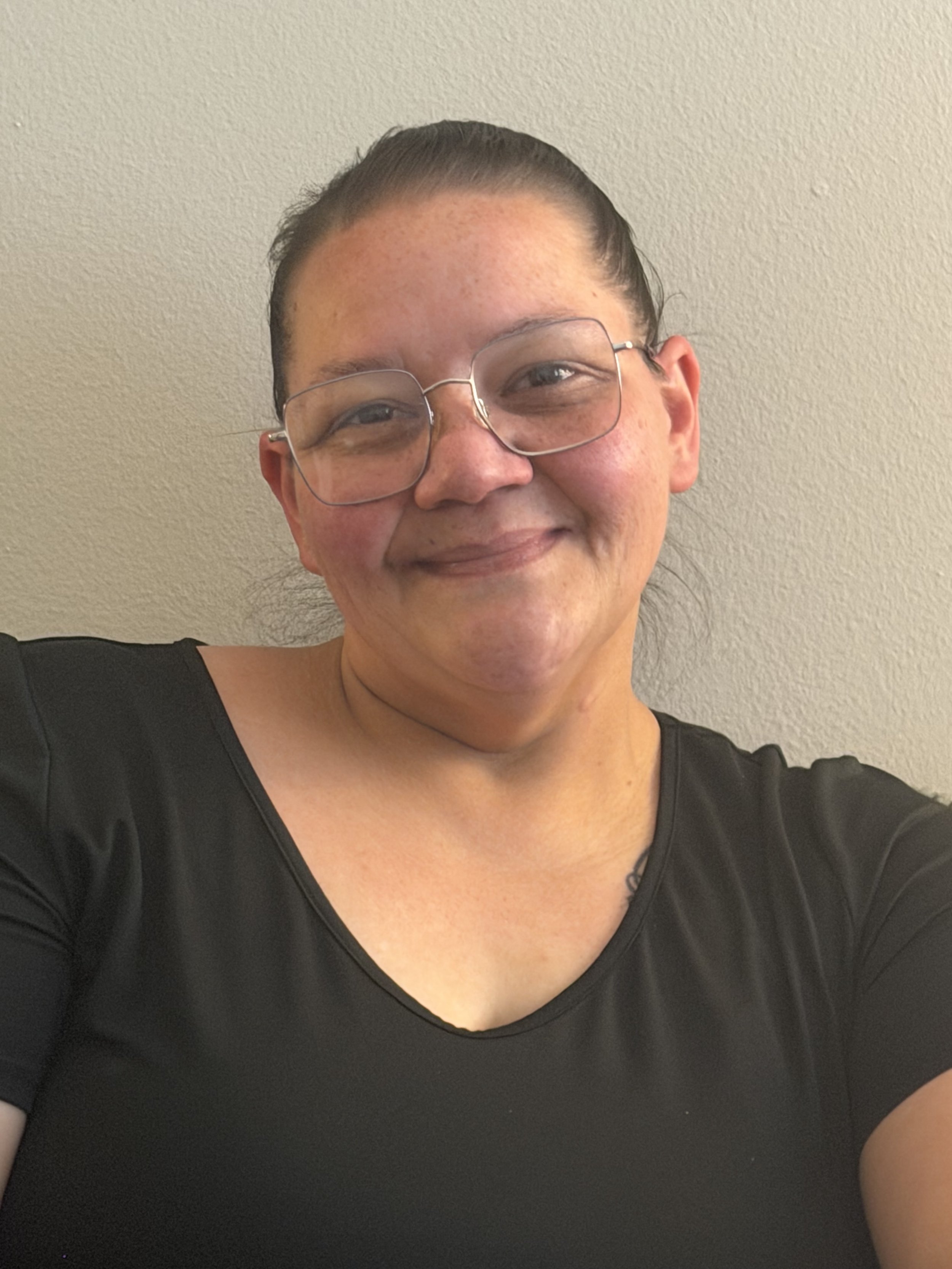 Close-up of a woman smiling, wearing glasses and a black top, against a plain wall background.