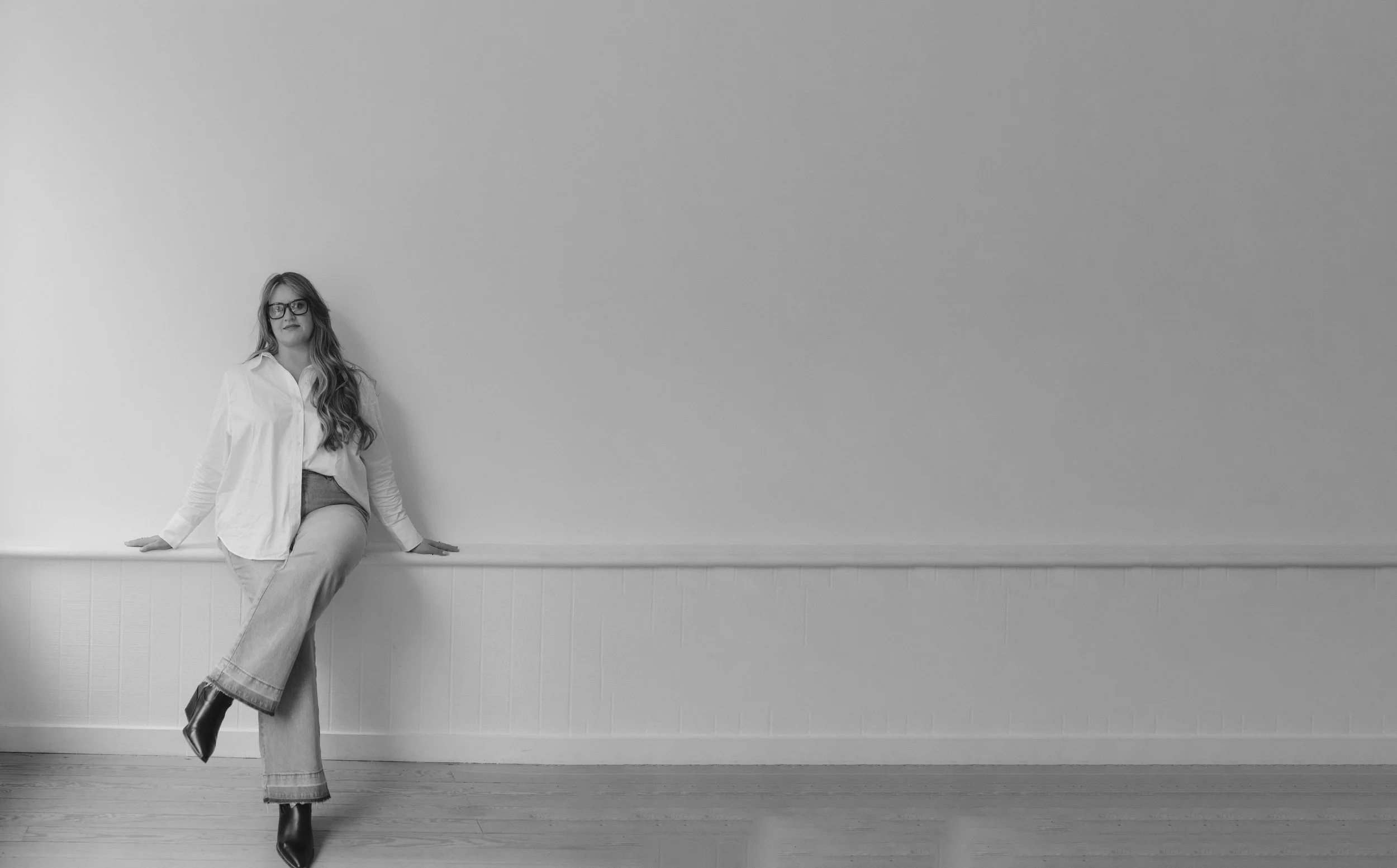 A woman with glasses and long wavy hair in a white shirt and jeans sitting on a ledge against a plain wall.