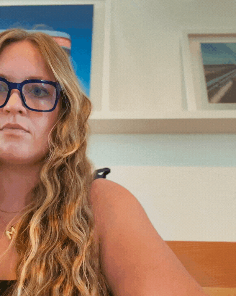 Close-up of a woman with long curly red hair and blue glasses, sitting in a room with a white wall and framed photos or artwork in the background.