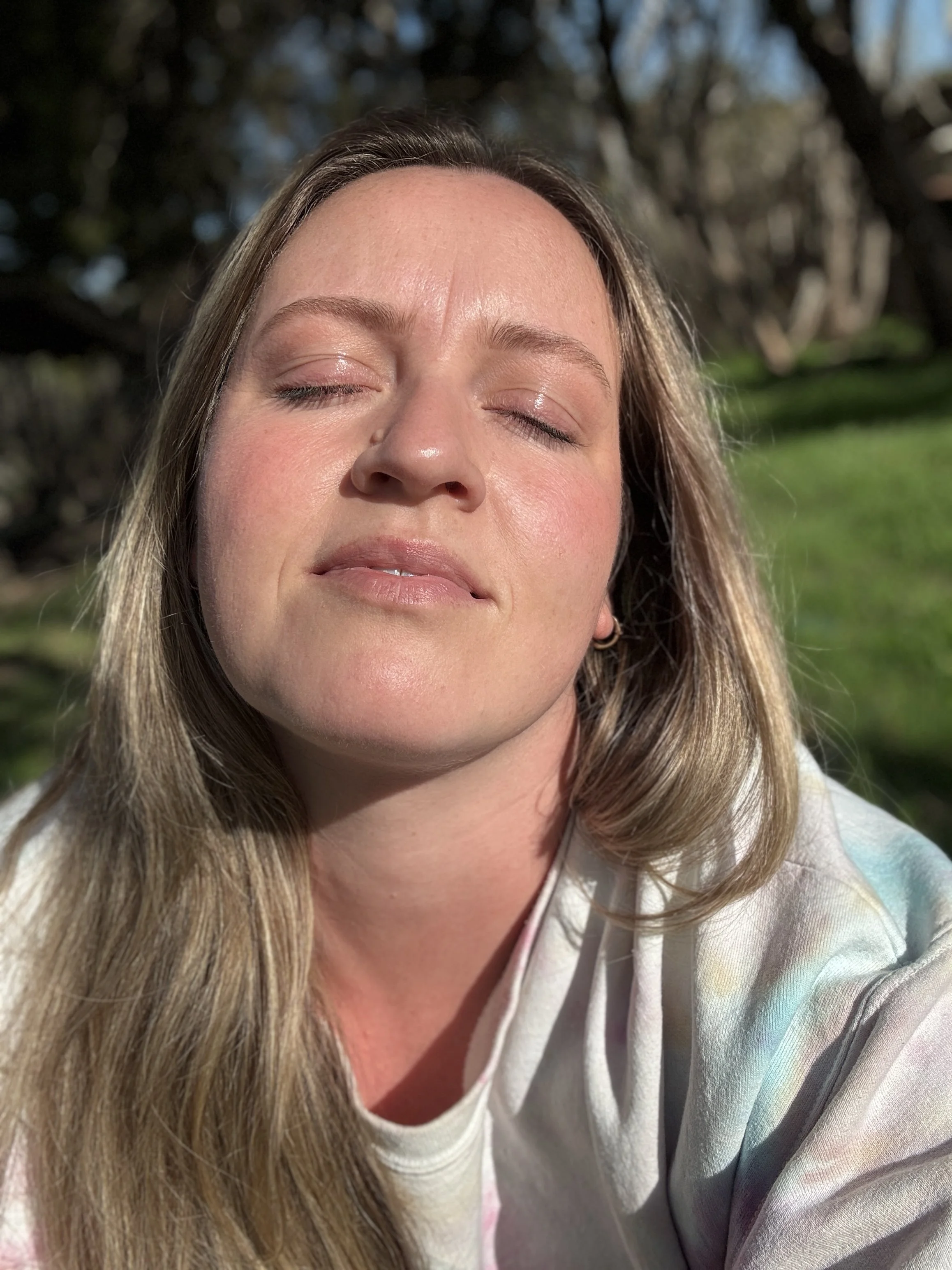 Close-up of a woman with long hair, eyes closed, smiling slightly, outdoors in sunlight with trees and grass in the background.