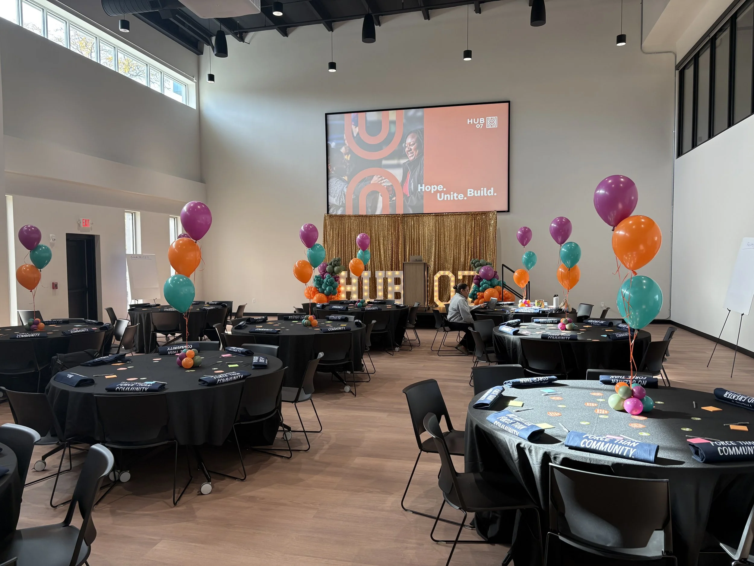 A large, modern conference room decorated for an event with balloon bouquets in purple, orange, and teal colors. Round tables are set with black tablecloths, blue napkins, and small pink notes. A big screen on the wall displays the words "Hope. Unite. Build." with a logo. There are people seated at some tables and a person walking near the decorations.