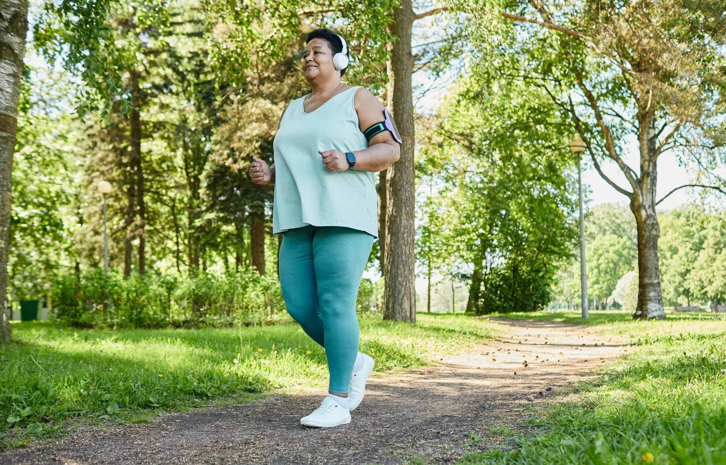 Woman jogging on a forest trail, wearing wireless headphones, a sleeveless top, turquoise leggings, white sneakers, and a fitness armband.