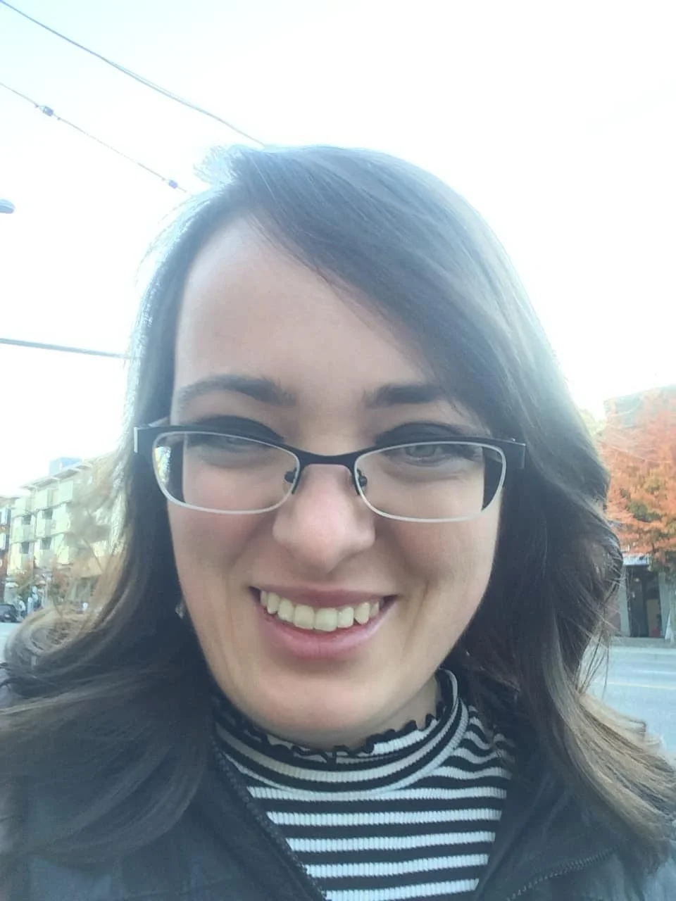 A smiling woman with glasses takes a selfie outdoors on a bright day, with buildings and colorful trees in the background.
