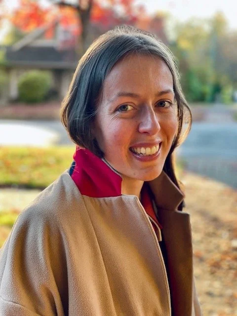 A young woman outdoors with fall foliage in the background, smiling and wearing a tan jacket with a red collar.