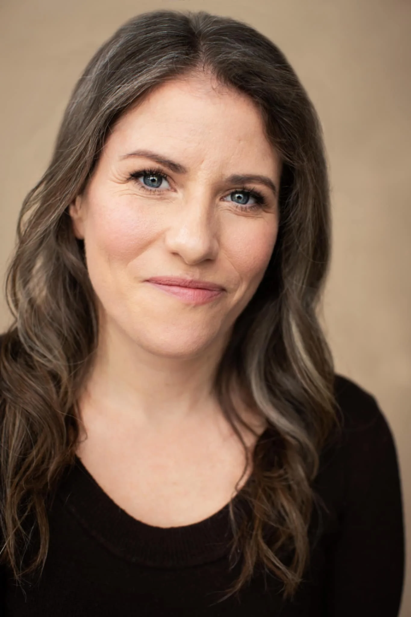 A woman with long wavy brown hair and blue eyes looking at the camera with a slight smile, wearing a black top against a neutral background.