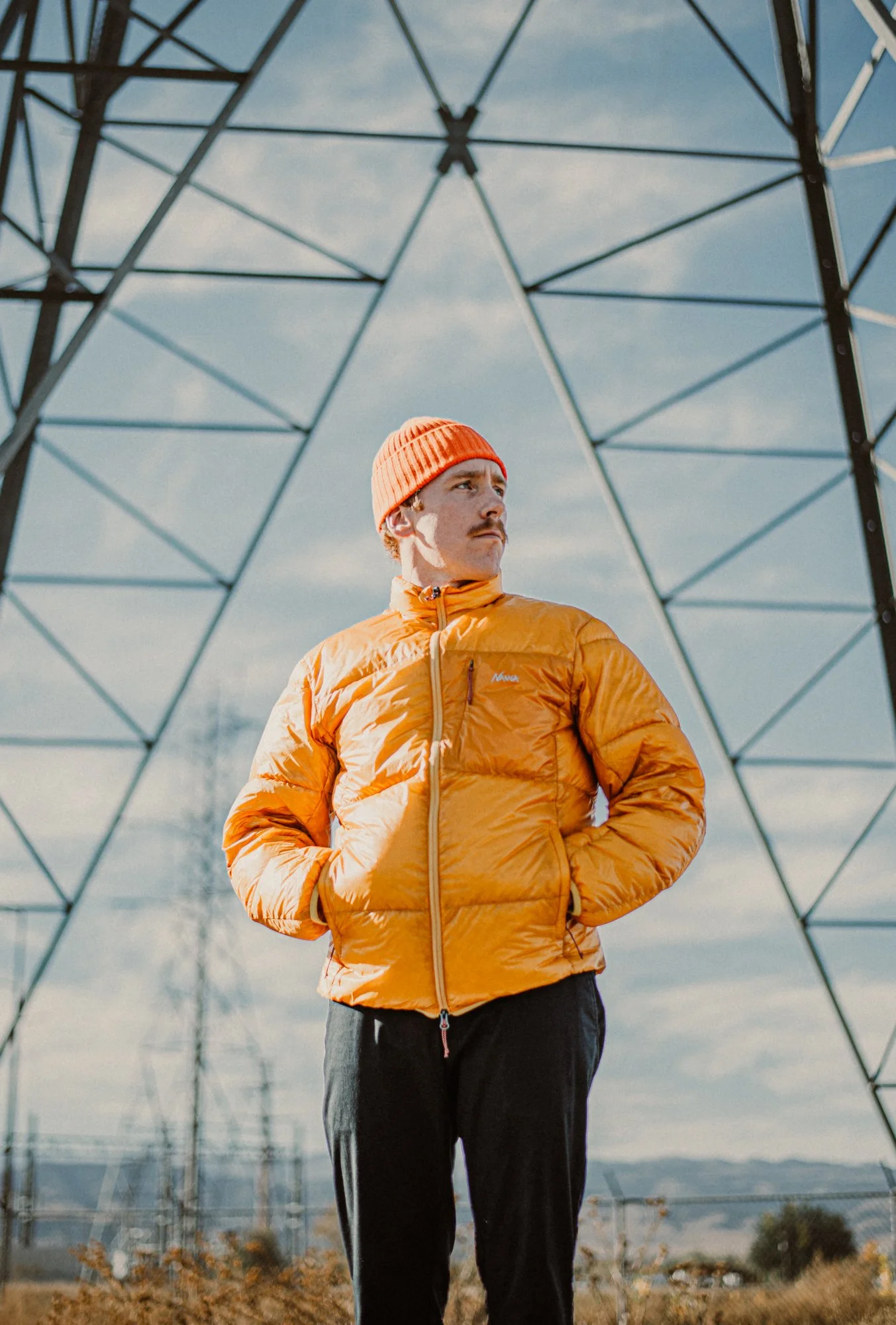Man in an orange jacket and orange beanie standing outdoors under a metal electrical pylon during daytime.