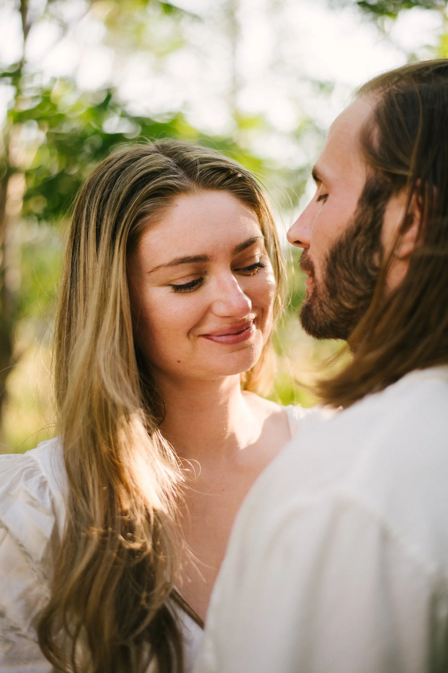 A woman and a man standing close together outdoors, smiling softly with their eyes closed, surrounded by blurred green foliage.