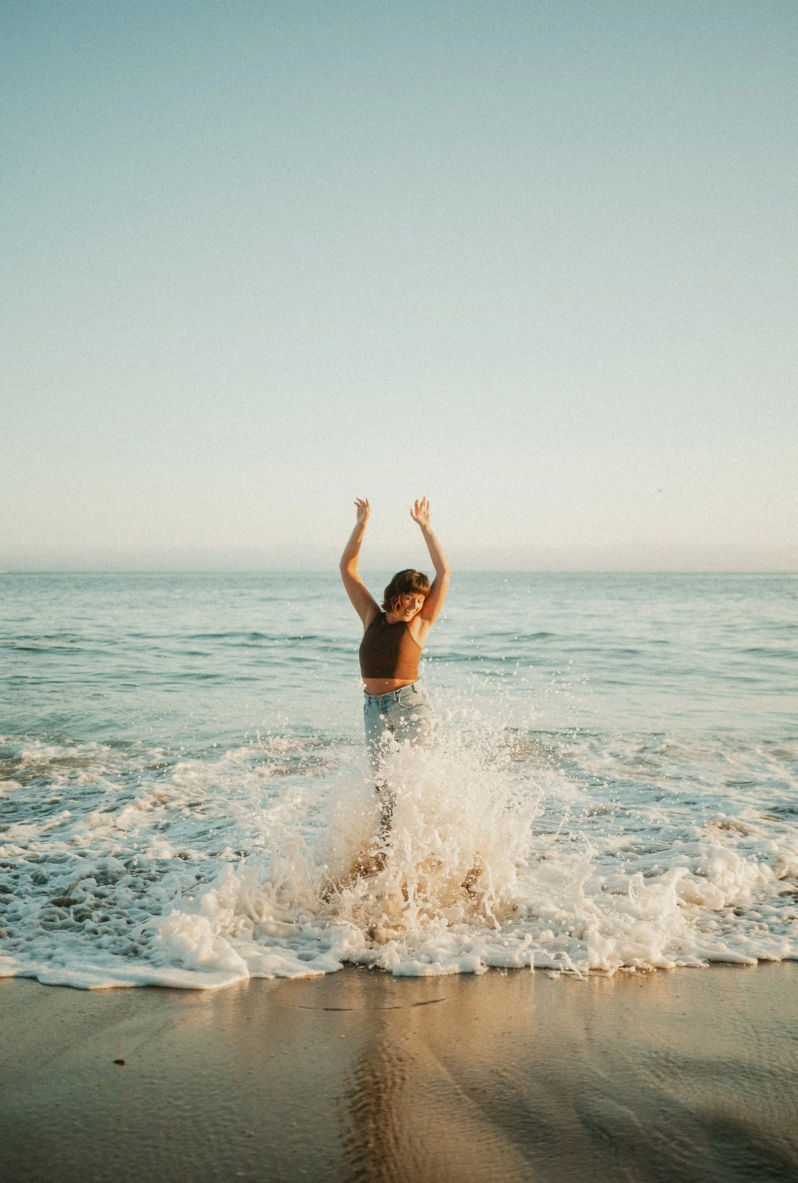 A woman standing in the ocean waves at the beach with her arms raised in the air, smiling.