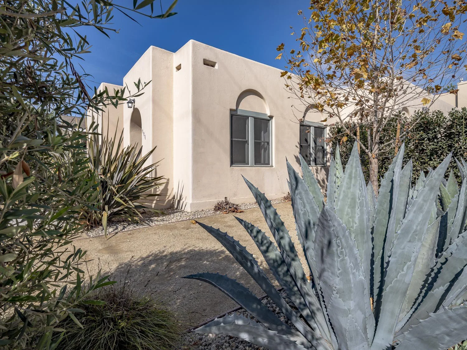 A beige stucco house with arched windows, surrounded by desert plants including a large agave and a small tree, under a clear blue sky.