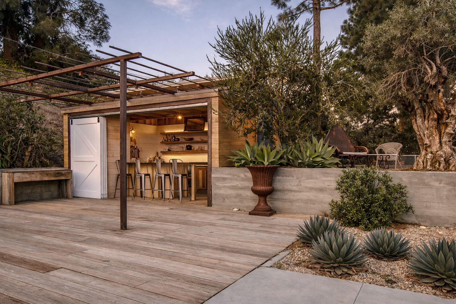 An outdoor patio area at dusk featuring a small wooden bar with stools, surrounded by plants including agave and a large tree, with outdoor seating and a gravel ground.