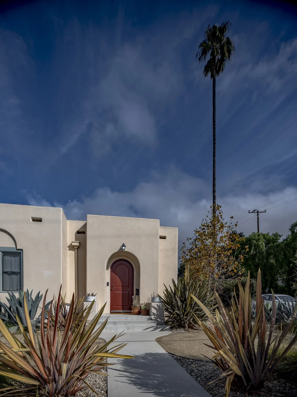A house with a red front door, surrounded by desert plants, under a blue sky with wispy clouds, with a tall palm tree in the background.