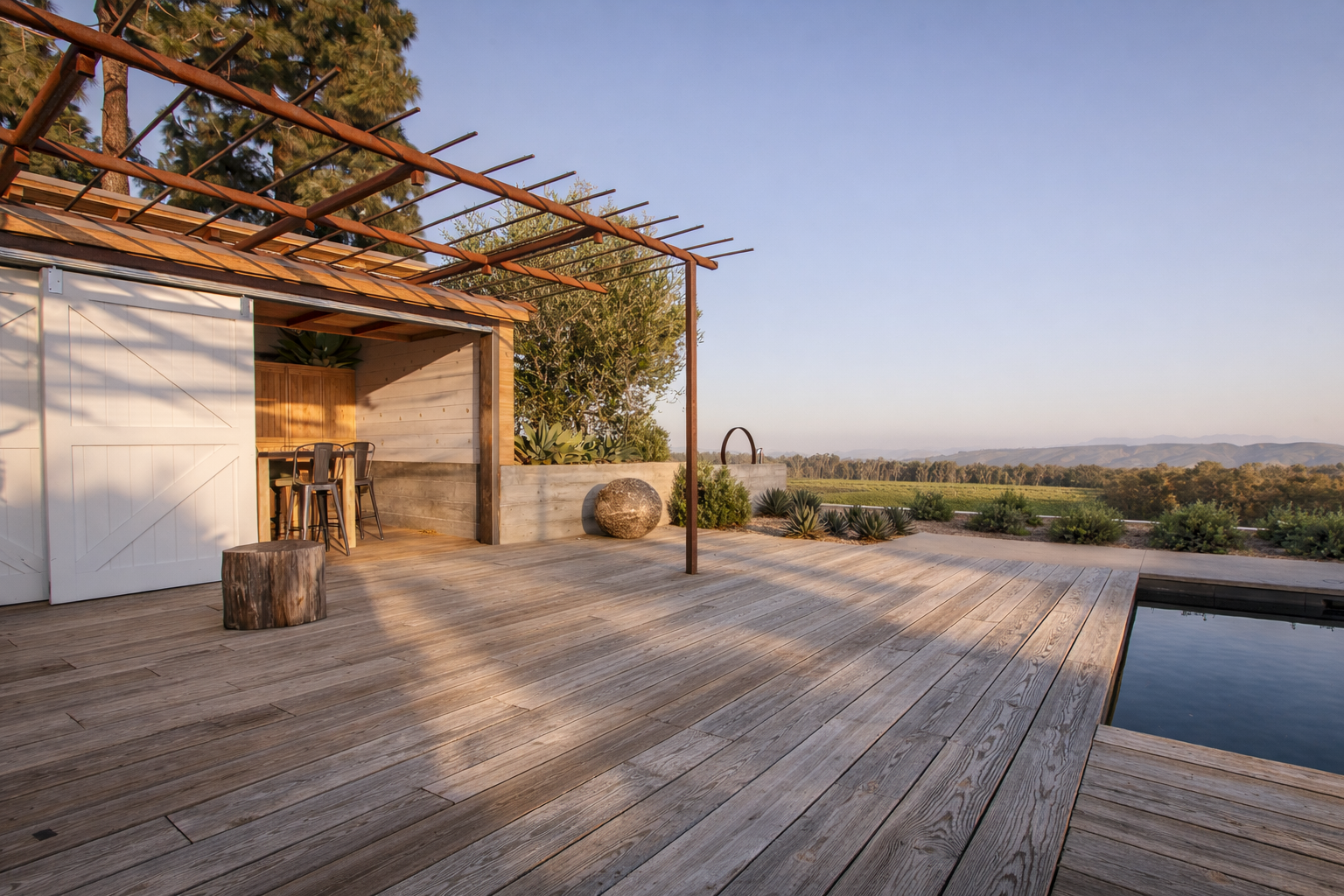 A wooden deck with a small pool, outdoor furniture, and surrounding landscape of trees and open fields in the distance under a clear sky.