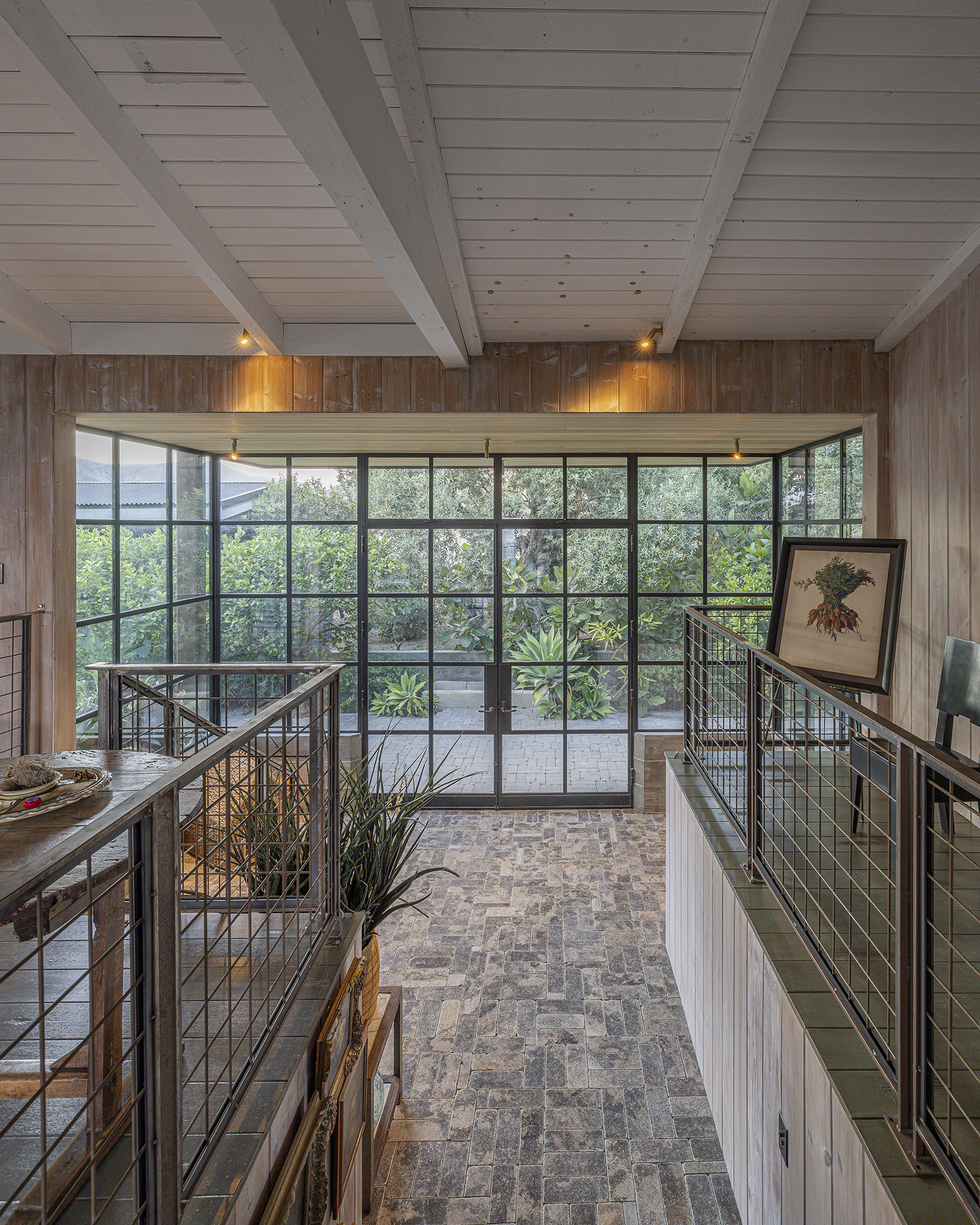 Interior view of a room with large glass windows, brick flooring, and wooden walls and ceiling. There are plants outside visible through the windows, and a framed picture leaning against the railing.