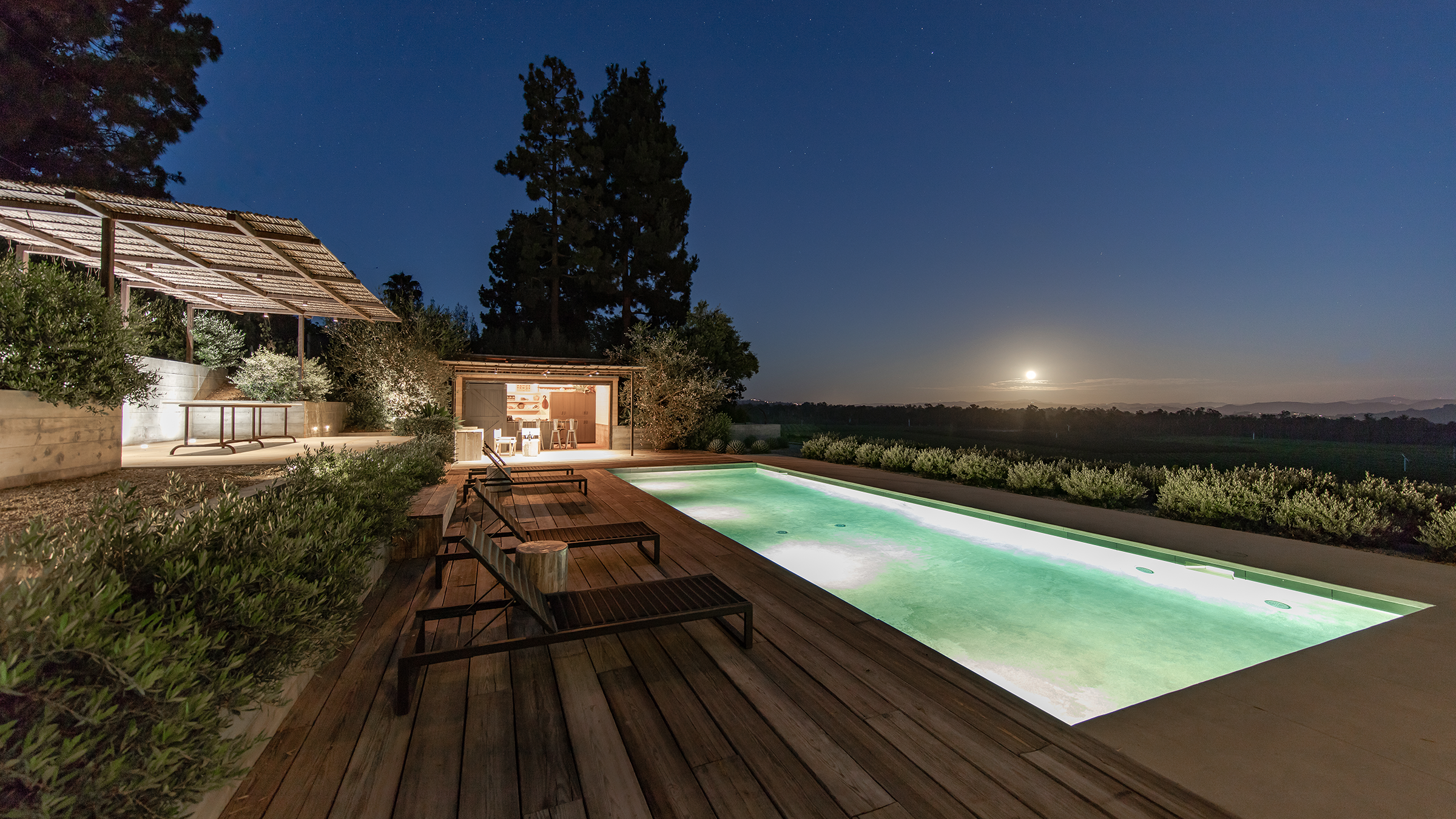 Night view of a backyard with a lit swimming pool, wooden deck, outdoor seating, a small indoor bar area, trees, and a full moon in the sky.