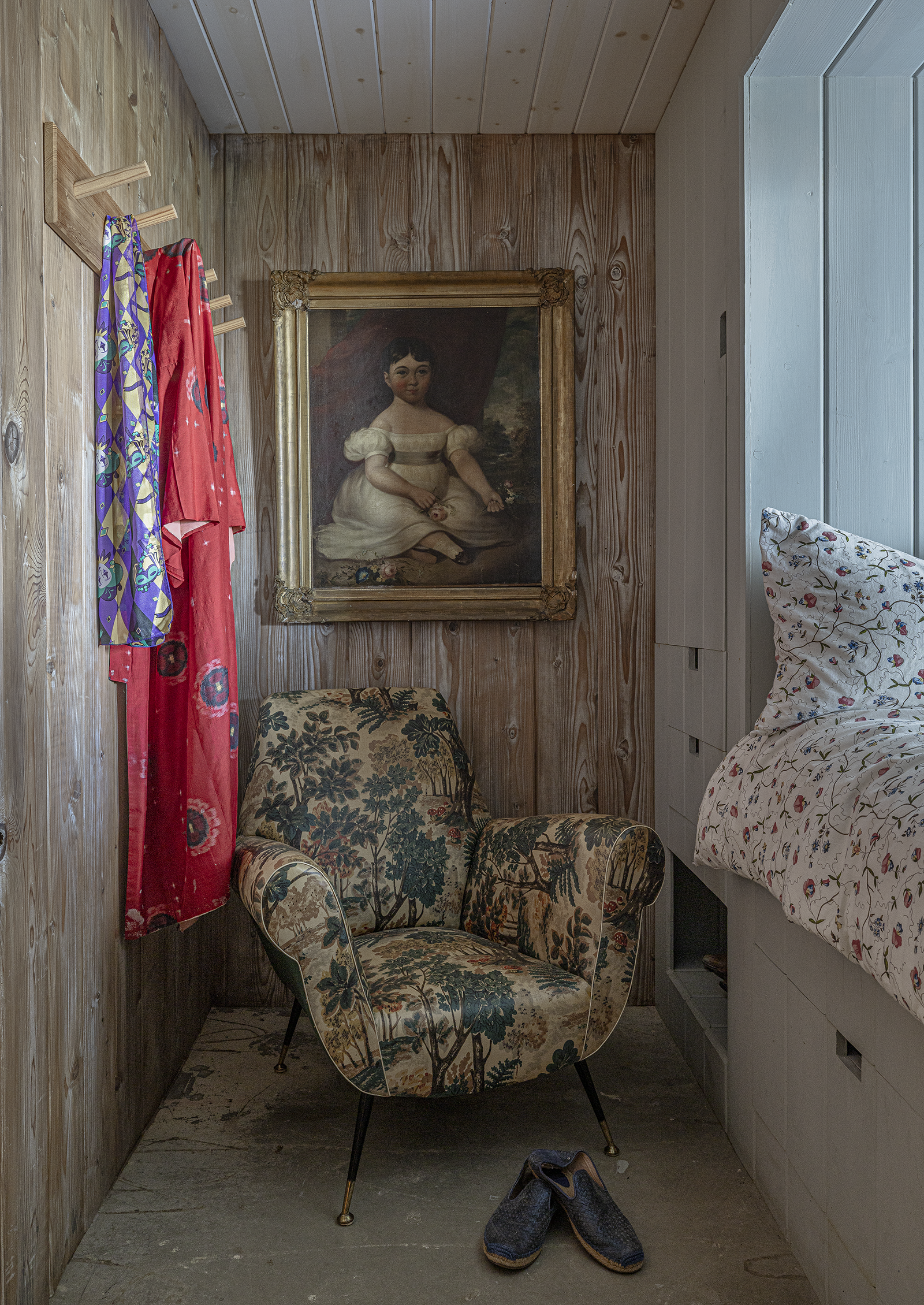 A cozy corner with a framed portrait of a young girl, a vintage armchair with floral upholstery, a pair of blue shoes on the floor, and a bed with a floral duvet in a room with wooden walls.